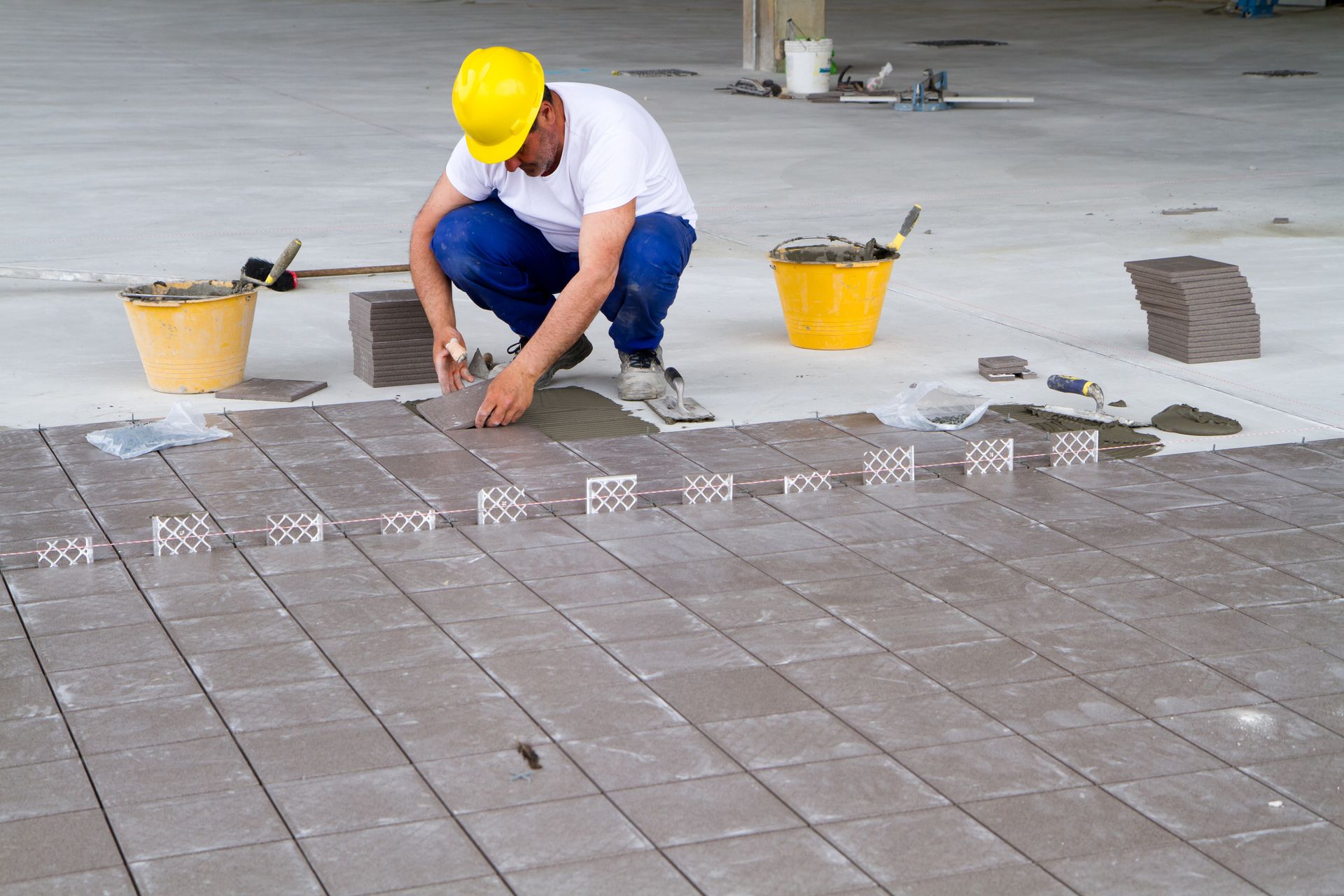 A man wearing a hard hat is laying tiles on a concrete floor.
