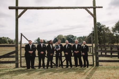 Seven people in suits standing under a large wooden arch outdoors on a grassy field