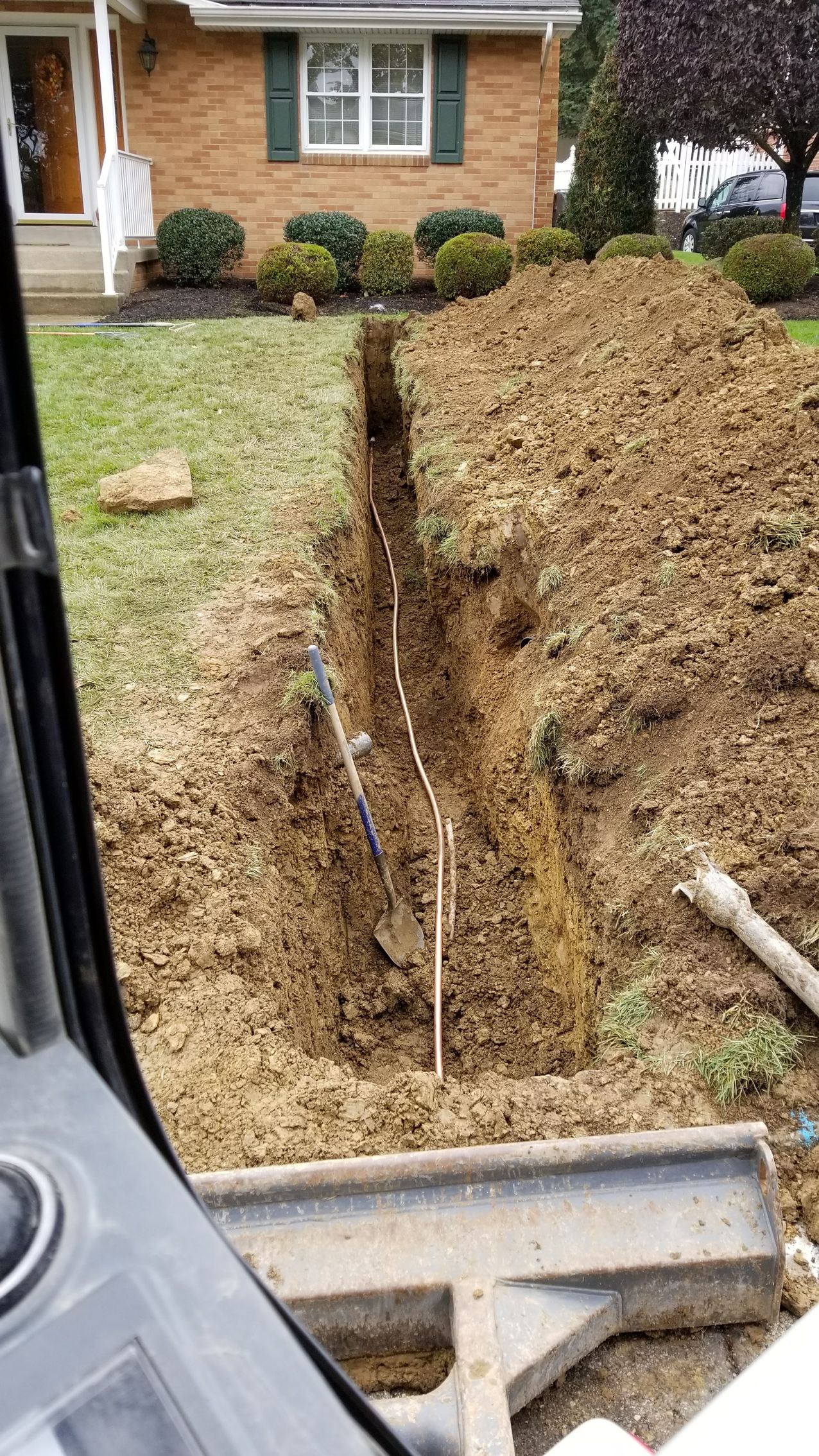 Trench dug in front yard reveals water pipes. Brown soil, green grass, and a brick house are visible.