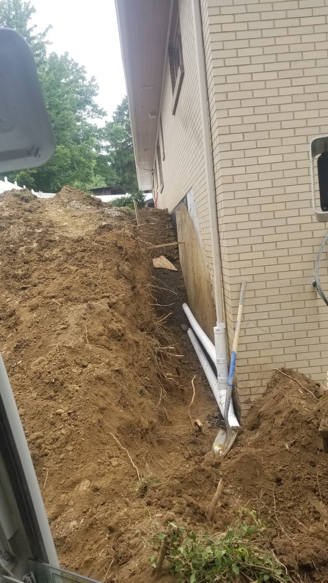 Trench dug along a brick building. Earth pile next to the wall. PVC pipes and tools are in the trench.