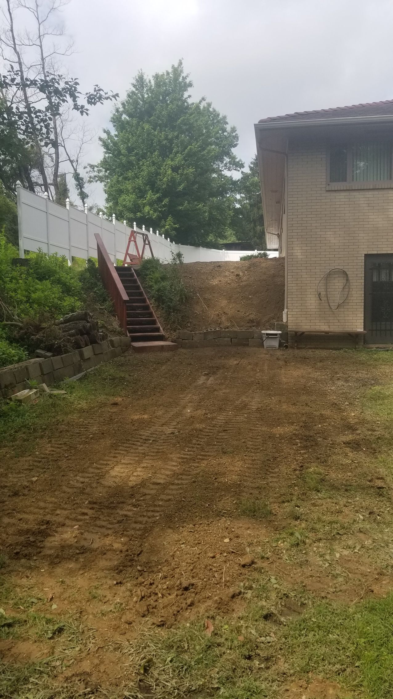 Backyard with mud, stairs leading up to a fence, and a house on the right. Overcast sky.