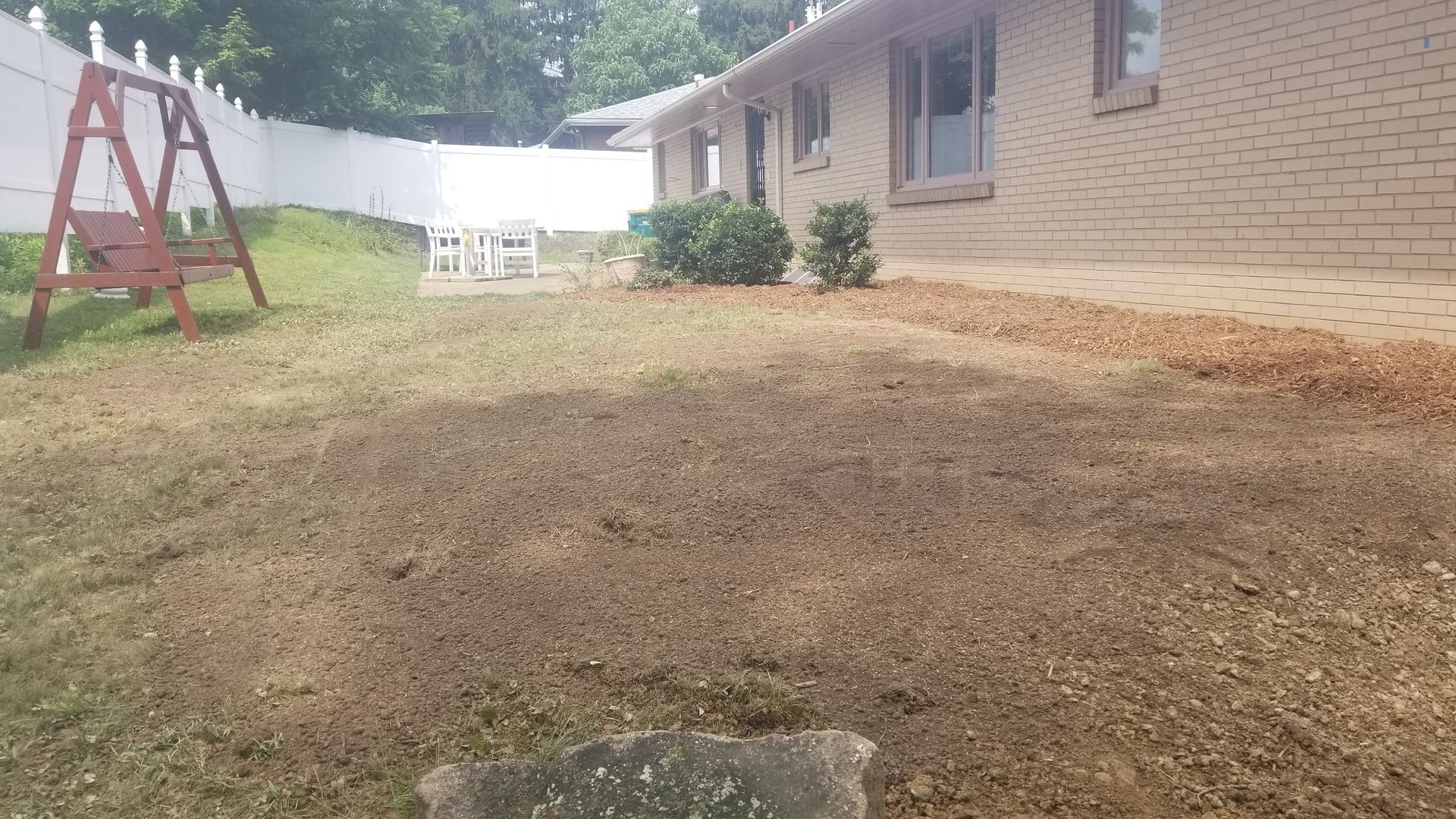 Yard with dead grass, brown gravel border, brick house, swing set, white fence.