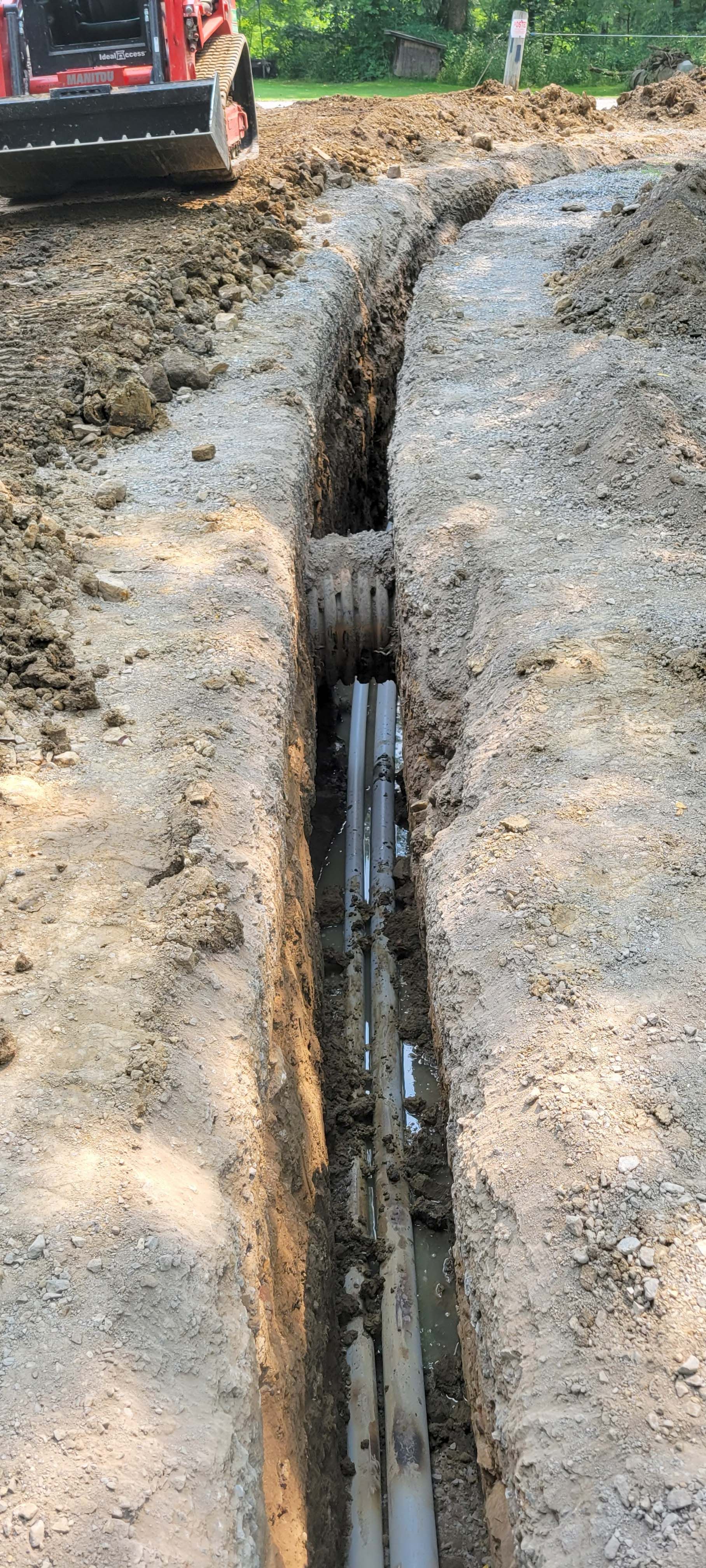 Trench dug in the ground, exposing underground pipes. A small red excavator is visible in the background.