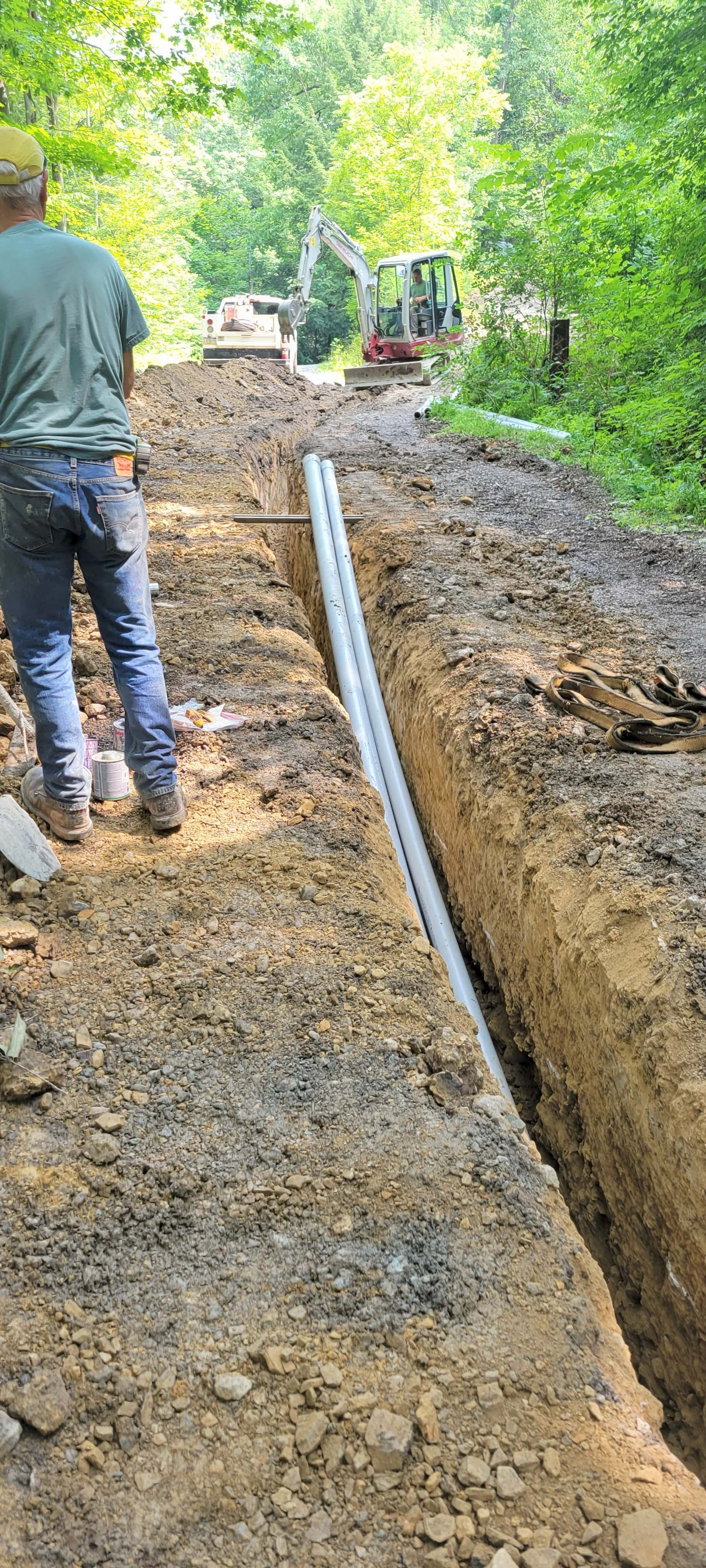 A person stands near a trench with white pipes, with construction equipment and trees in the background.