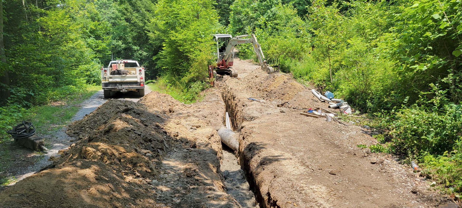 A dirt road is being excavated with heavy machinery, a truck is parked in the distance. Lush trees frame the scene.