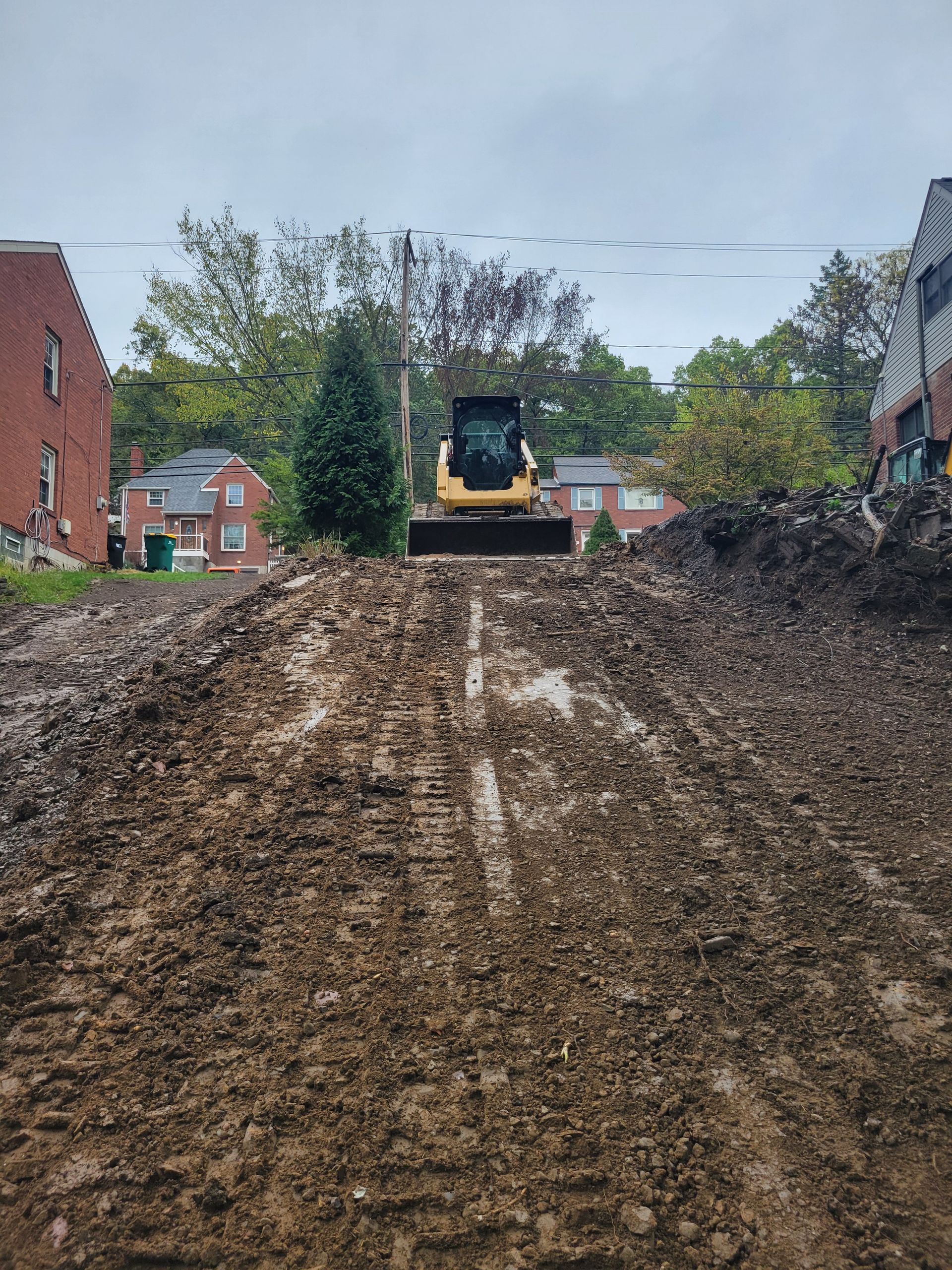 Yellow excavator on a mound of dirt with residential buildings in the background. Cloudy sky.