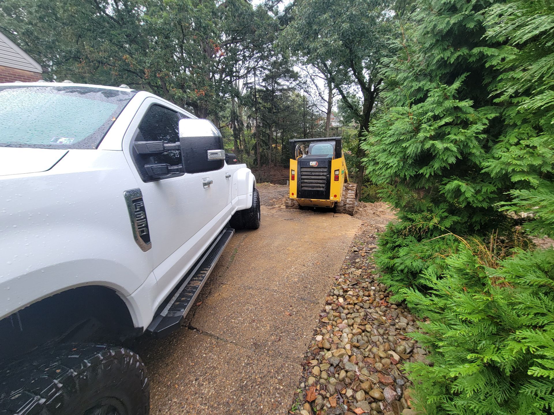 White truck and yellow skid steer on gravel driveway; greenery on the right.