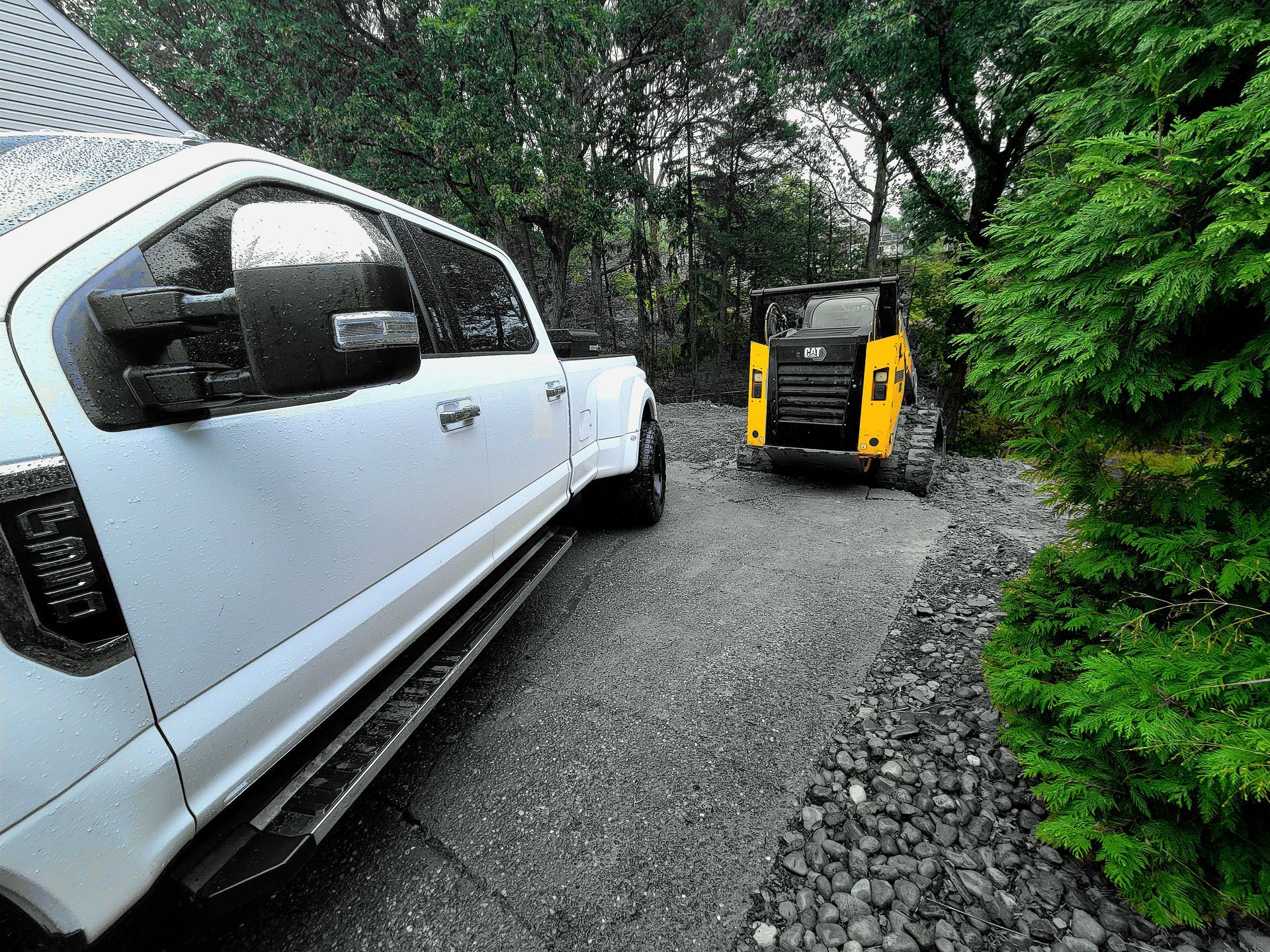 White truck parked on gravel driveway next to a yellow skid steer loader in a wooded setting.