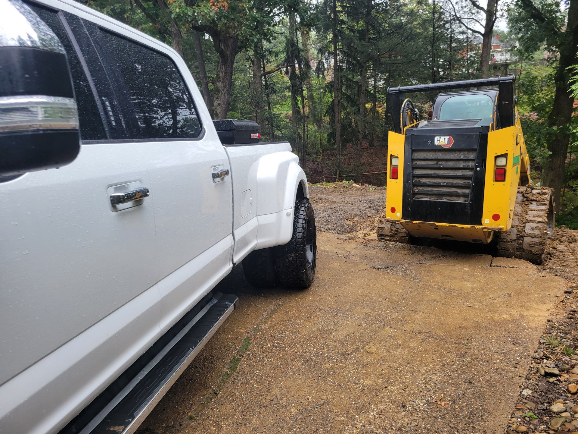 White pickup truck parked next to a yellow skid steer on a gravel driveway in a wooded area.