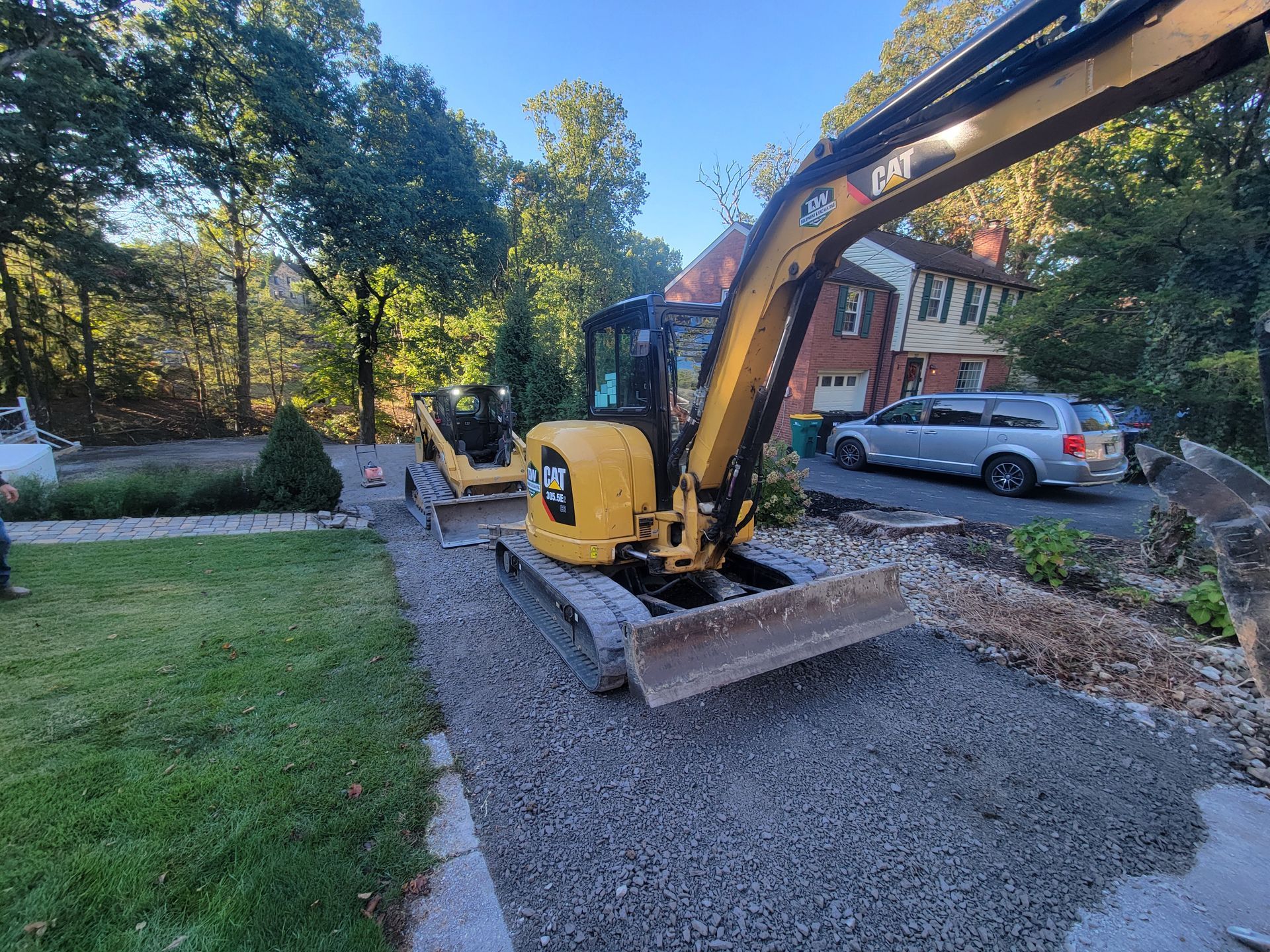 Yellow CAT excavator on a gravel path near a house, trees, and a parked car.