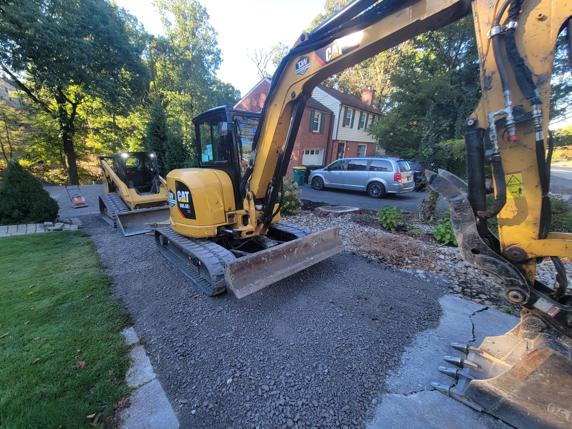 Yellow excavator on gravel, digging near a house, with a blue sky background.