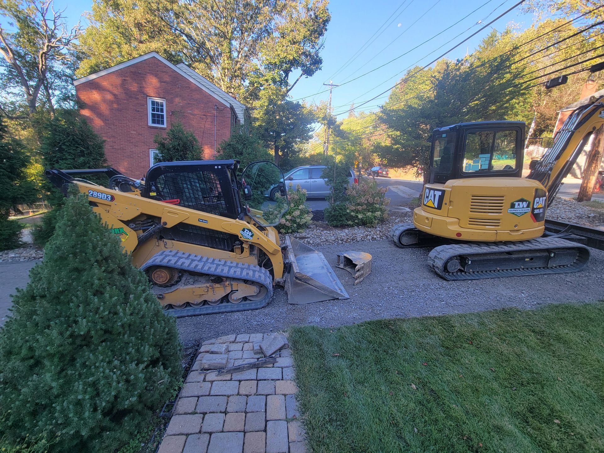 Two yellow construction vehicles on a gravel driveway. One is a skid steer, the other an excavator. Trees and a brick building are in the background.