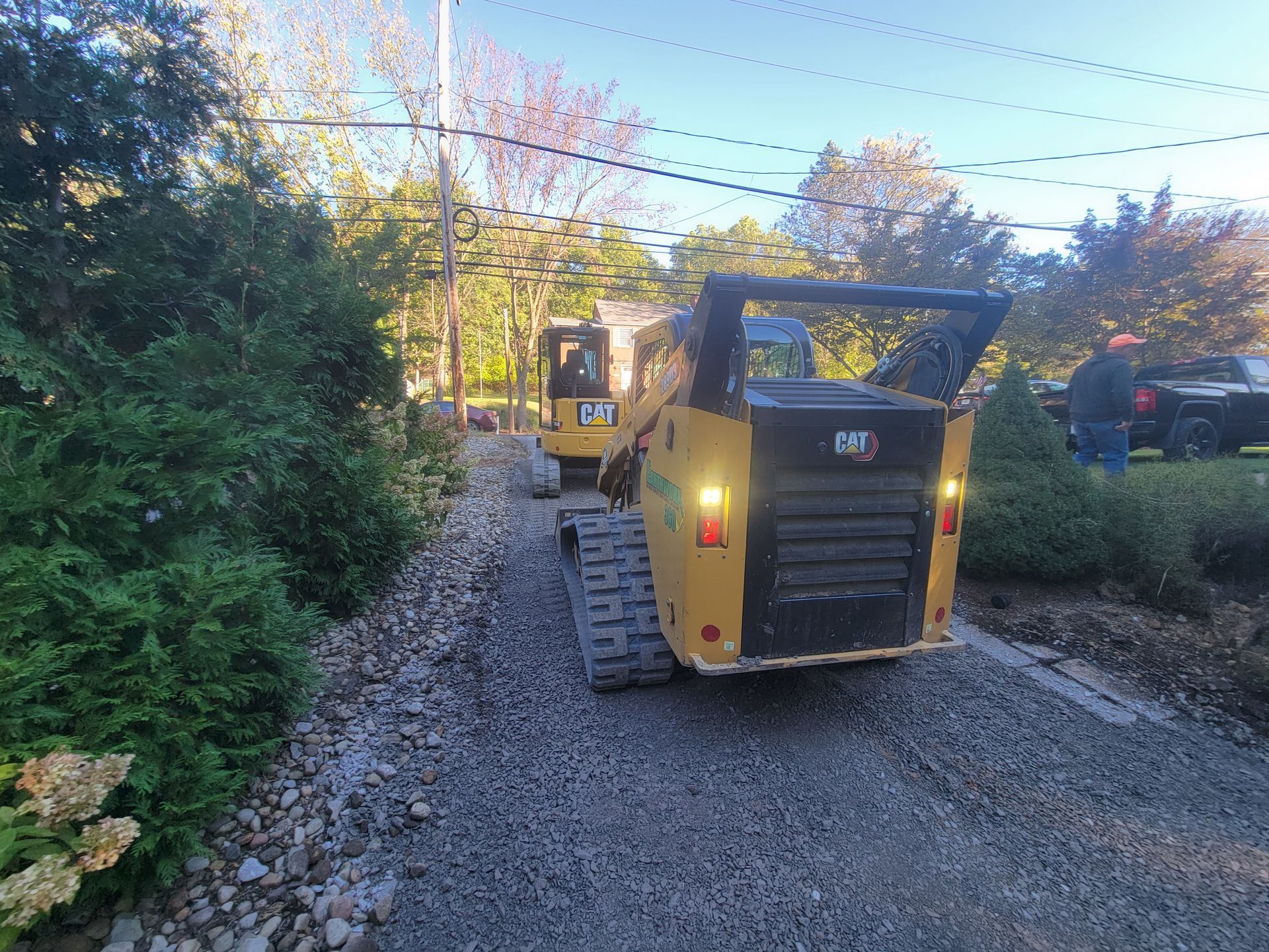 Two yellow skid steer loaders on a gravel driveway, preparing or working on the surface. Trees and house in the background.