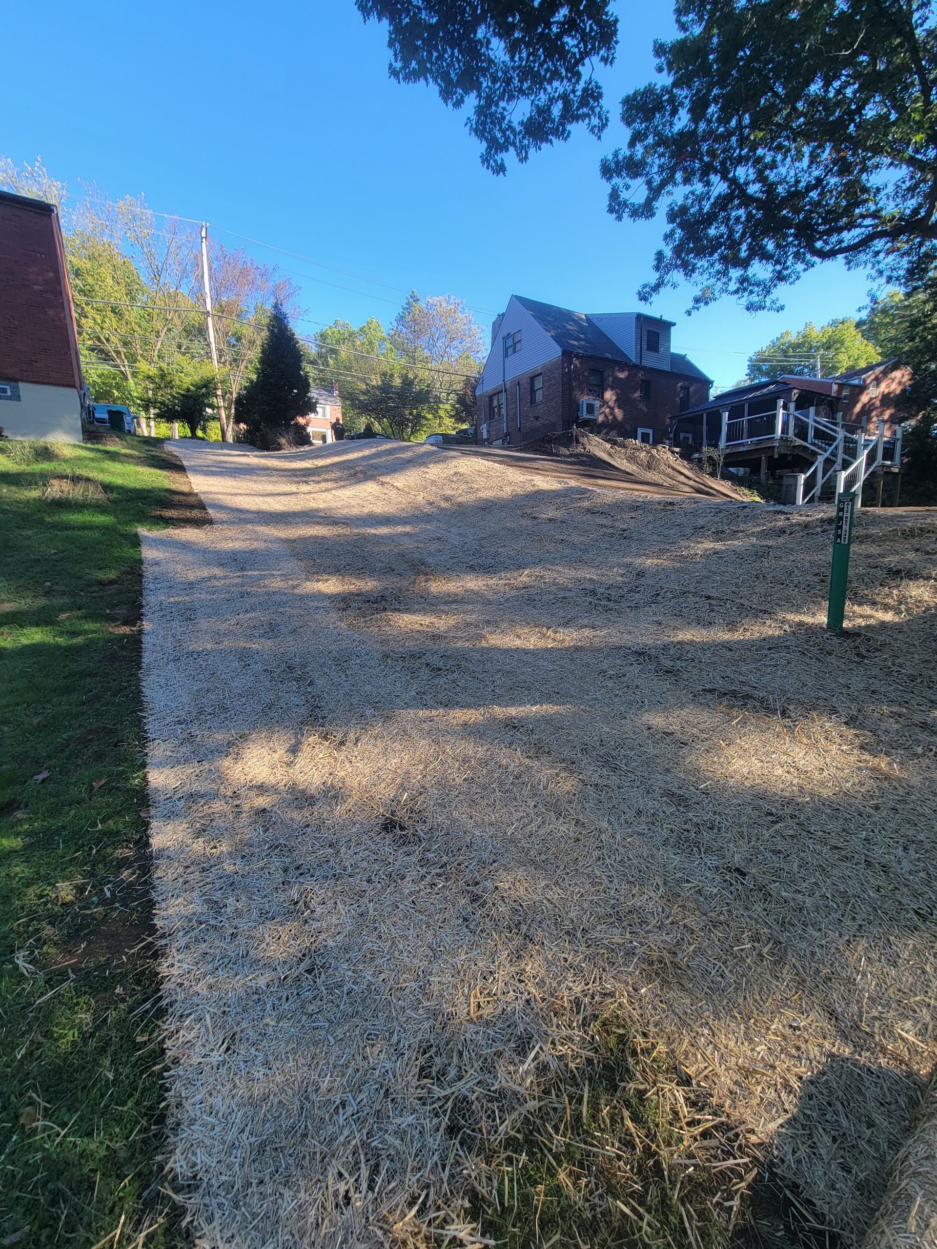 Gravel-covered slope in a yard, leading toward houses under a blue sky, with trees and grass on either side.