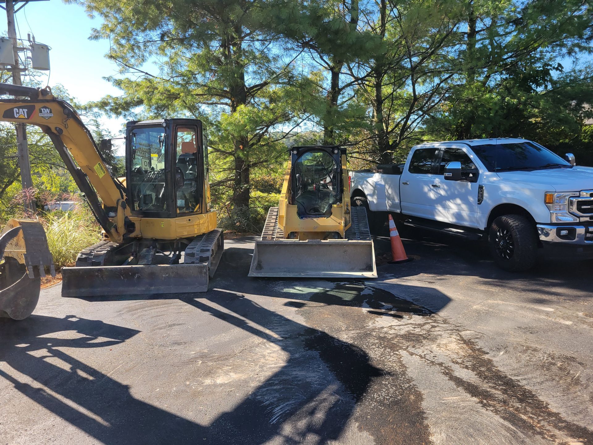 Yellow excavator and skid steer next to a white pickup truck on asphalt with a pothole.
