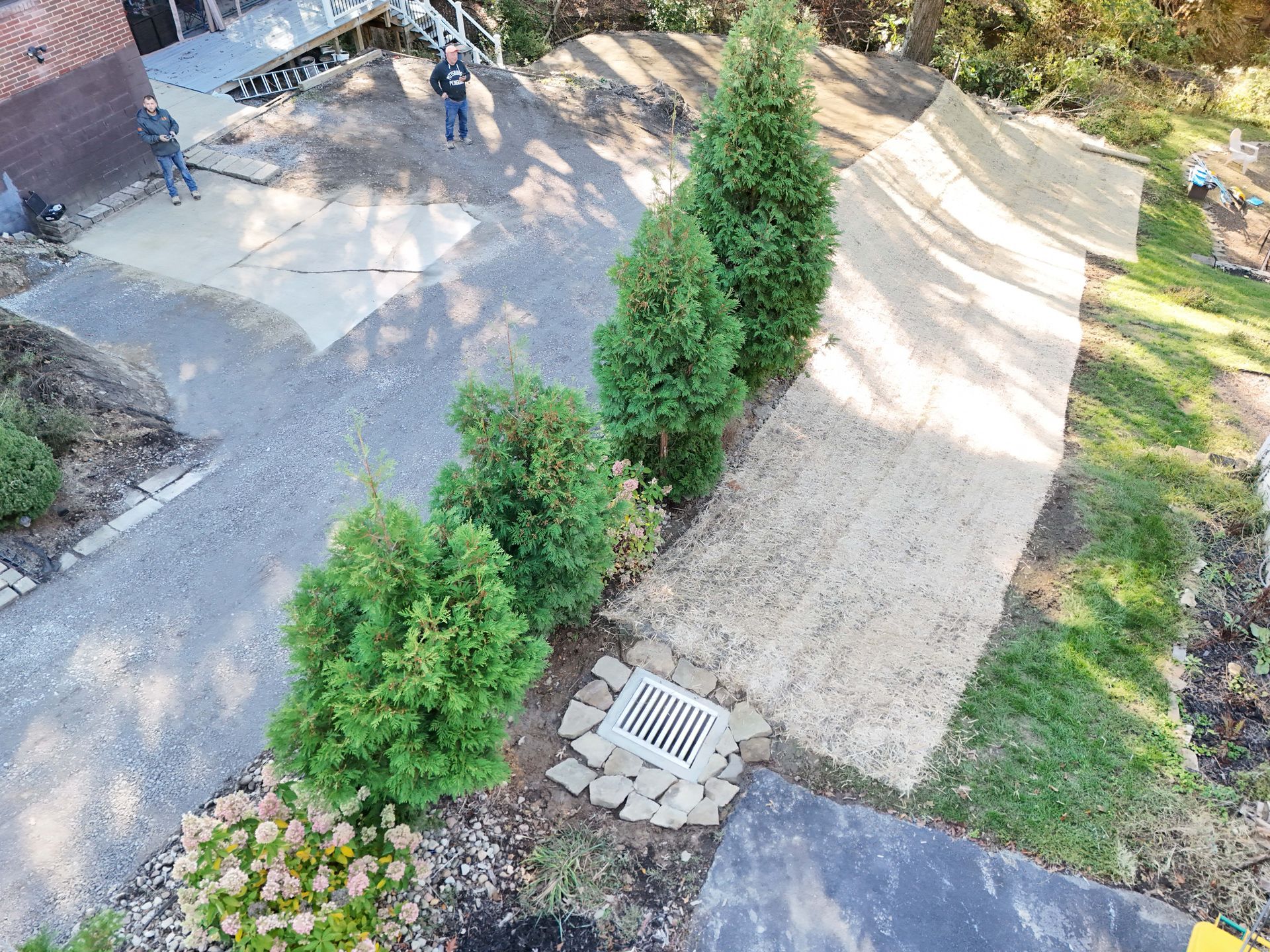An overhead view of a driveway with landscaping, including trees and a drain. Two people stand on the driveway.