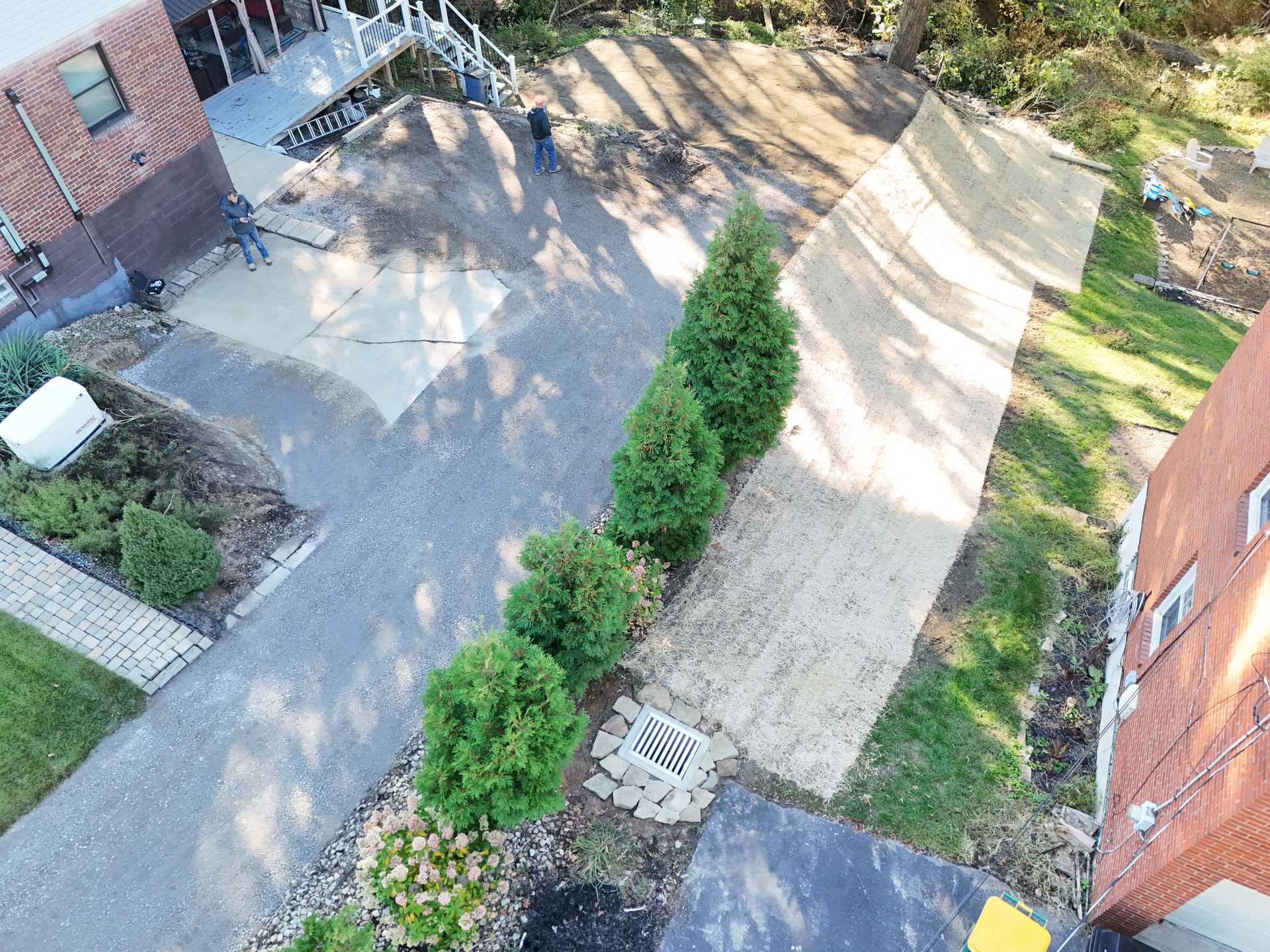 Overhead view of a paved driveway and lawn with a brick building. Several small trees are in a row.