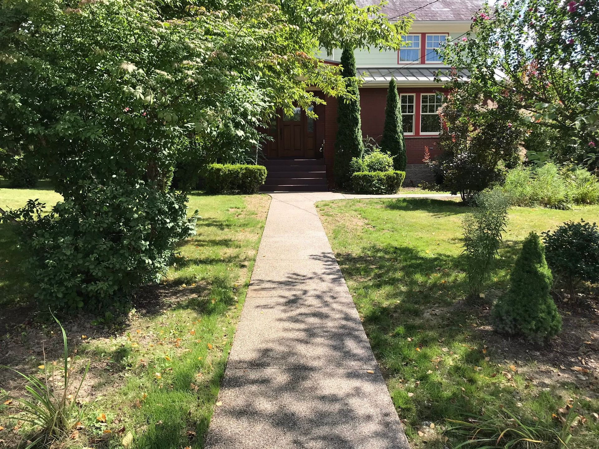 Concrete walkway leads to a house with a red brick facade, surrounded by trees and green grass.