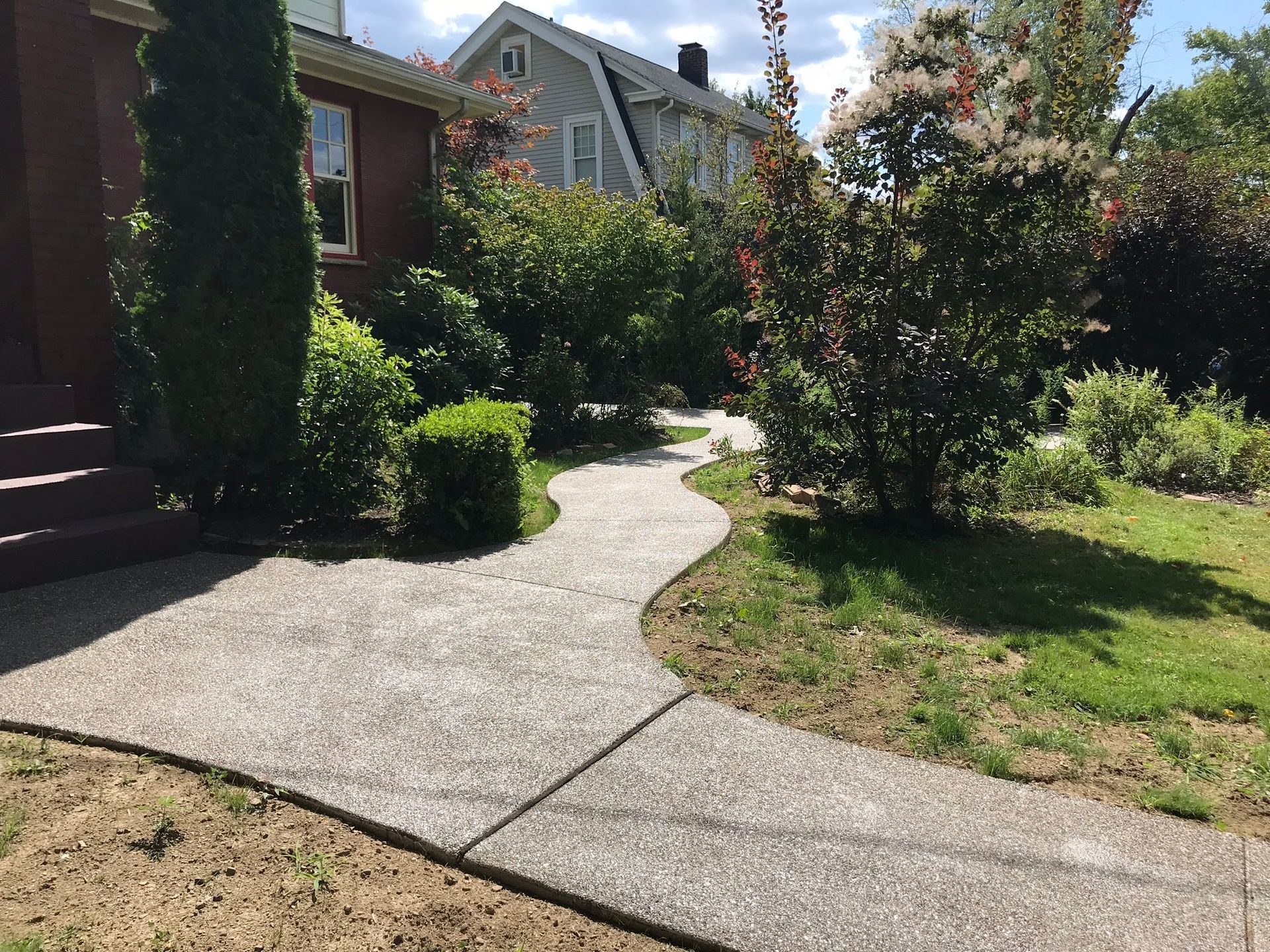 Concrete path curves through a garden with green bushes, leading to a house with red brick.