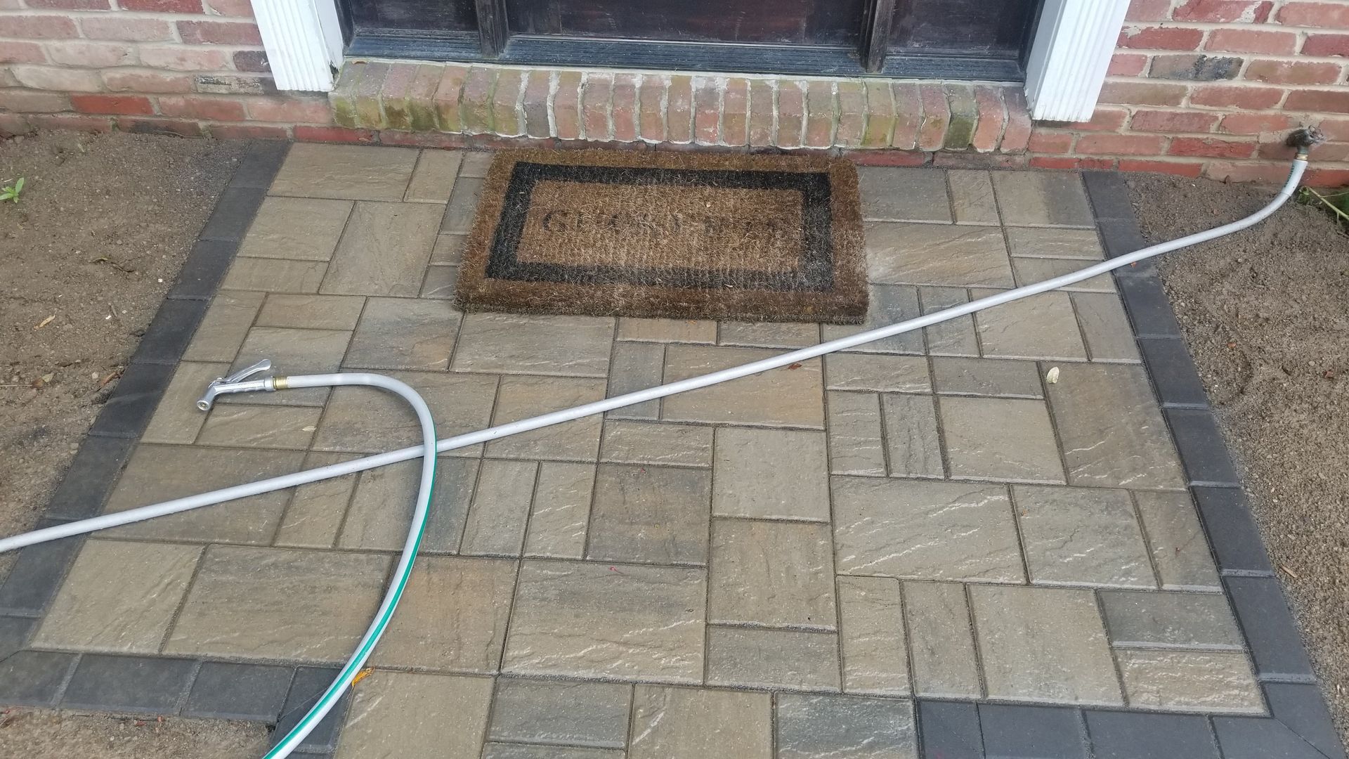 A hose on a brick walkway leading to a front door with a welcome mat.