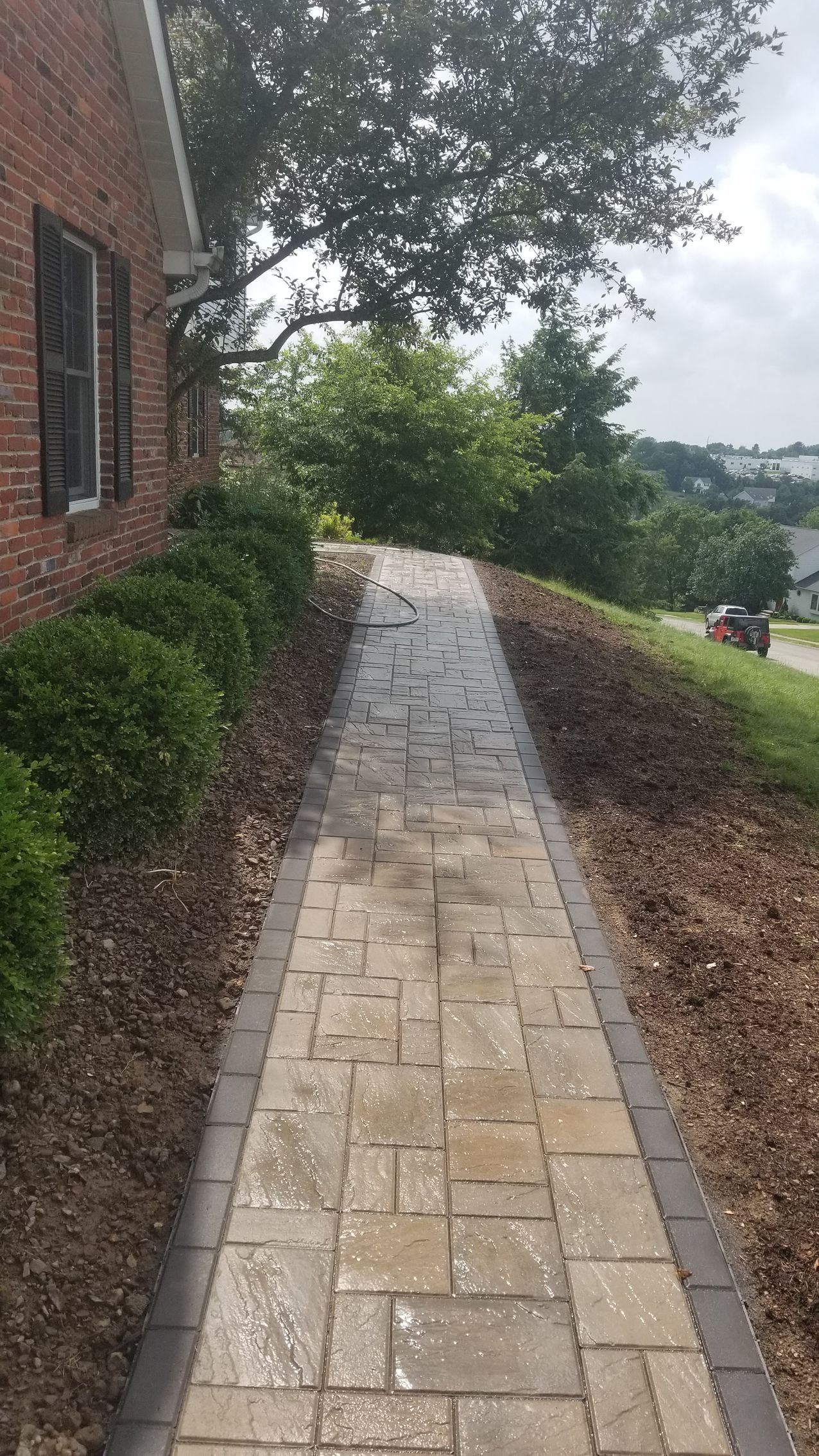 Brick walkway leading past brick building, green bushes, and hillside.