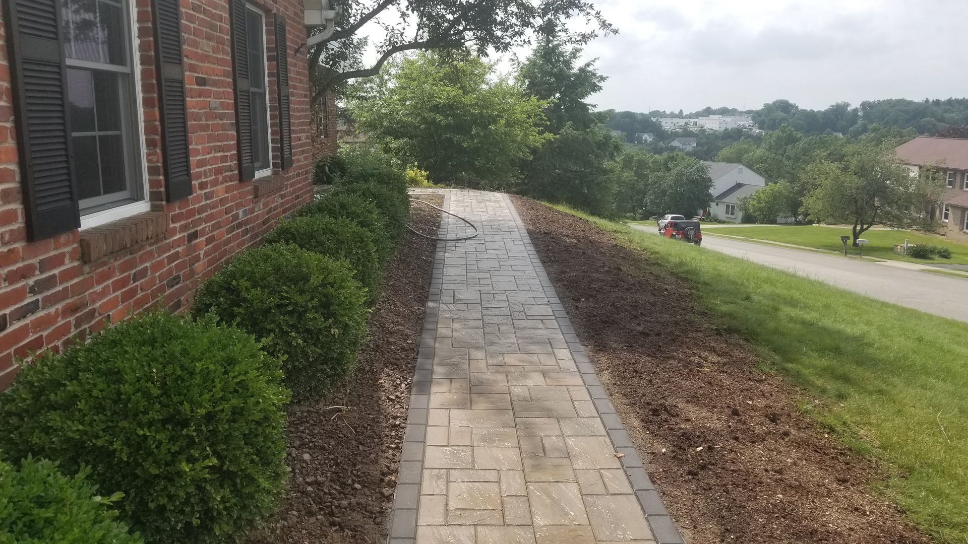 Brick home with walkway, shrubs, and mulch on a sloping hillside with houses in the distance.