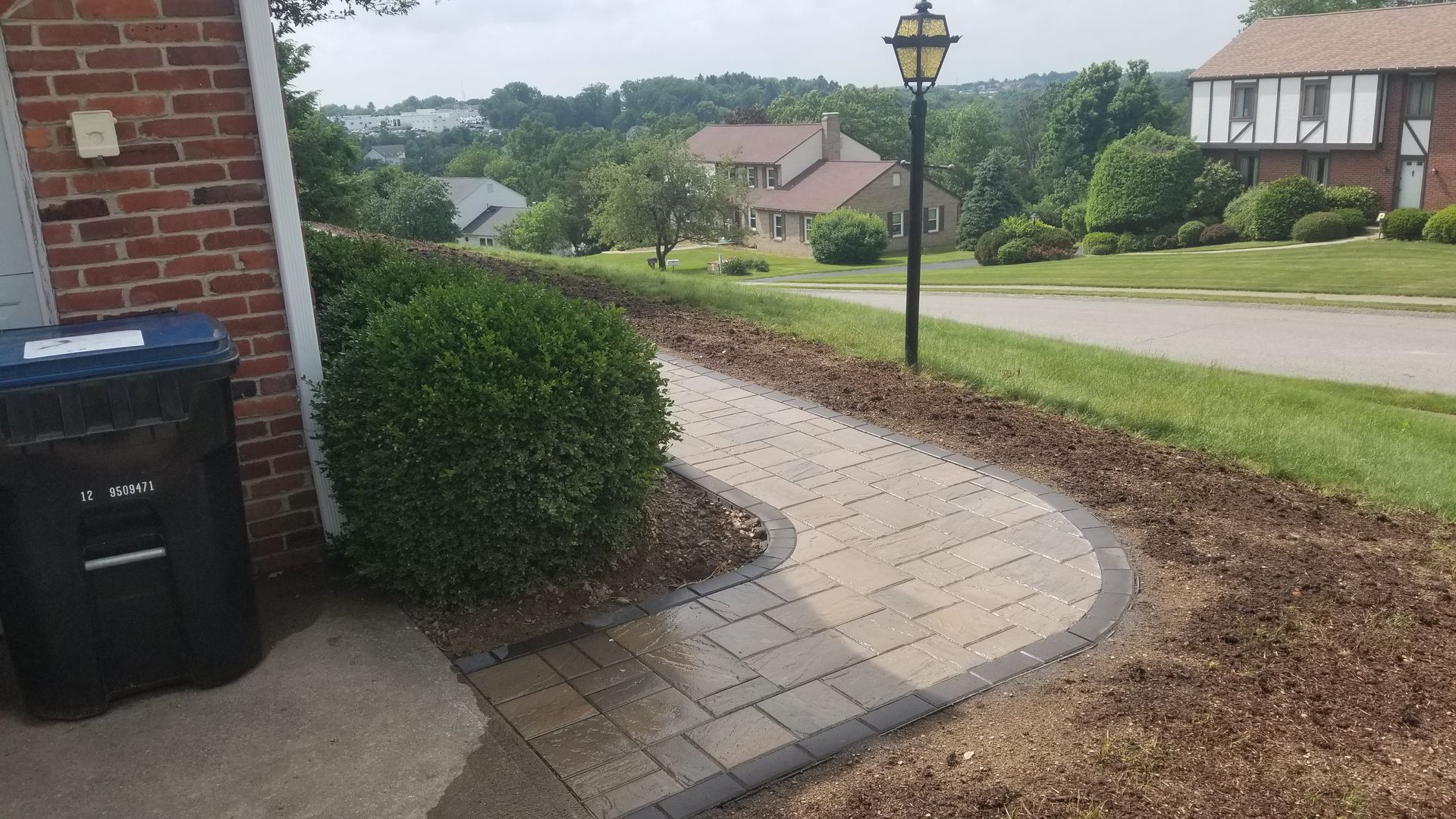 Brick pathway curves from a brick house to a street with green grass and trees in the background.