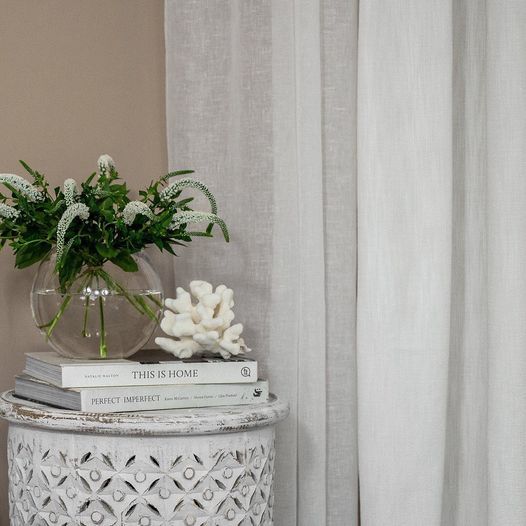 White curtains beside a carved, white side table with books, vase of flowers, and coral.
