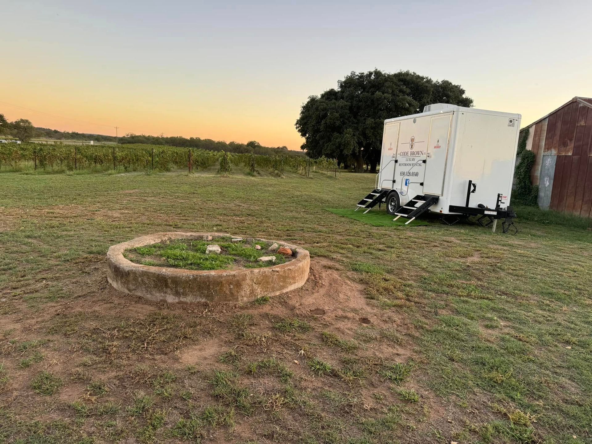 Grassy field at sunset with a circular stone fire pit and a white trailer beside a red shed.