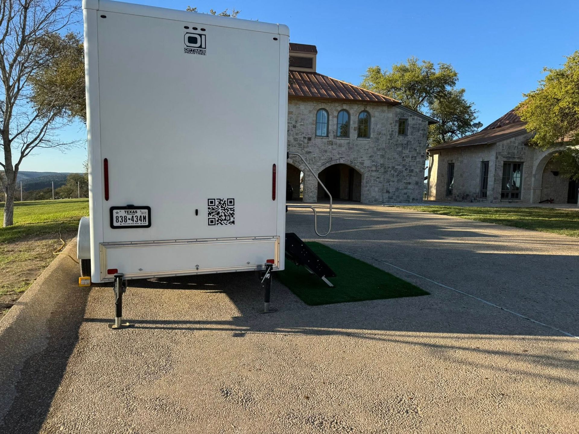White trailer parked on gravel beside a stone building with arched windows, with a green ramp at the entrance.