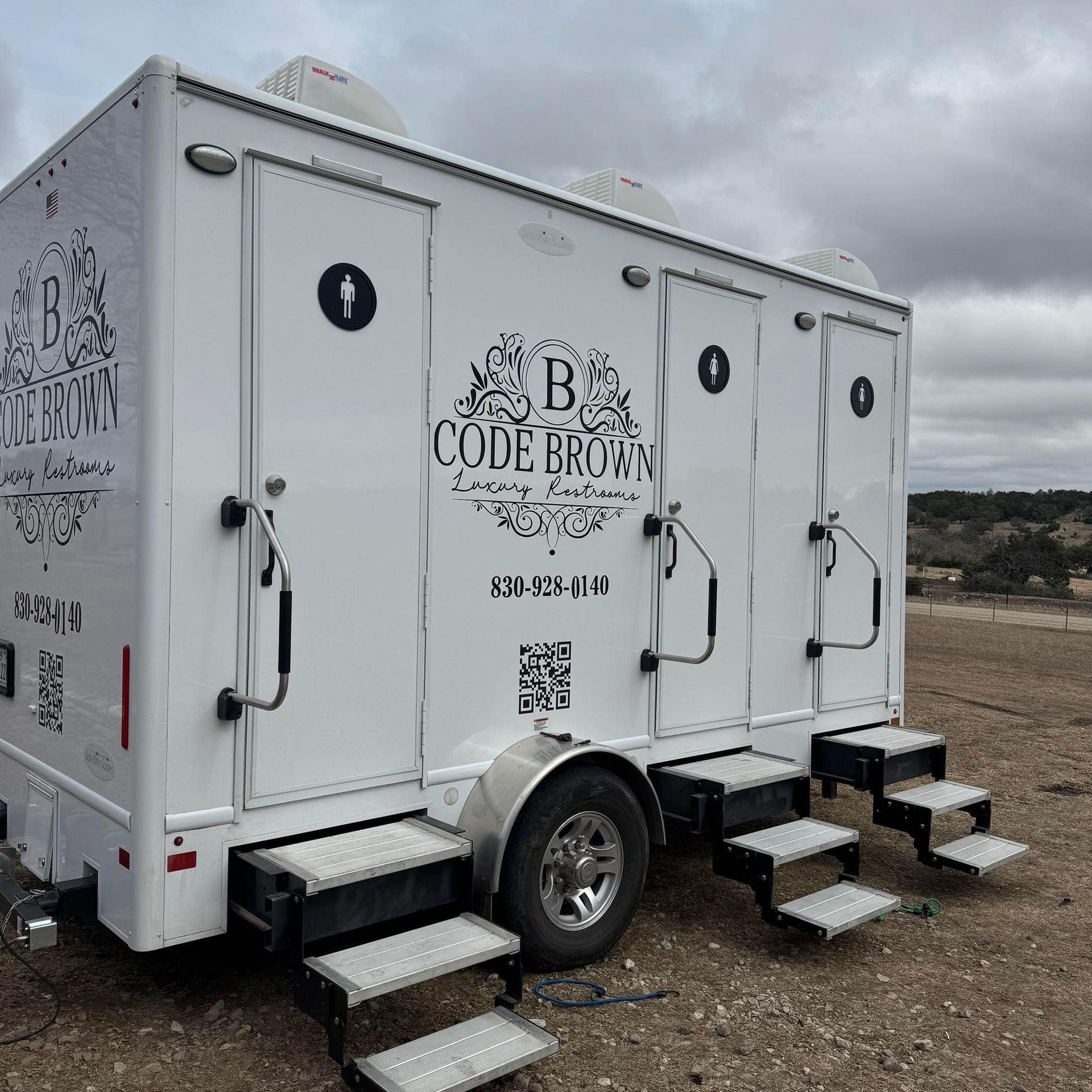 White mobile restroom trailer with steps, parked on a gravel lot under a cloudy sky.