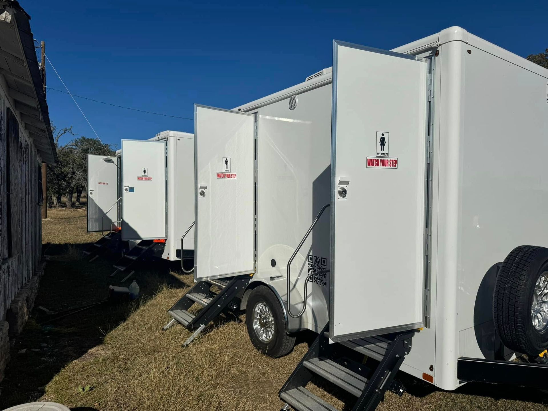 White portable restroom trailers lined up outdoors on grass under a clear blue sky.