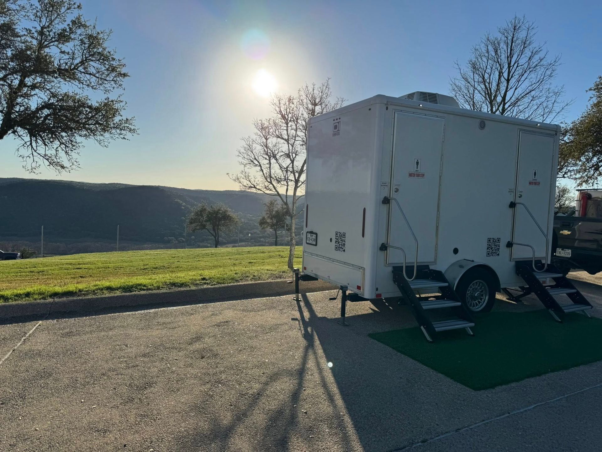 White portable trailer unit on a green mat in a sunny outdoor parking area with trees and hills in the background