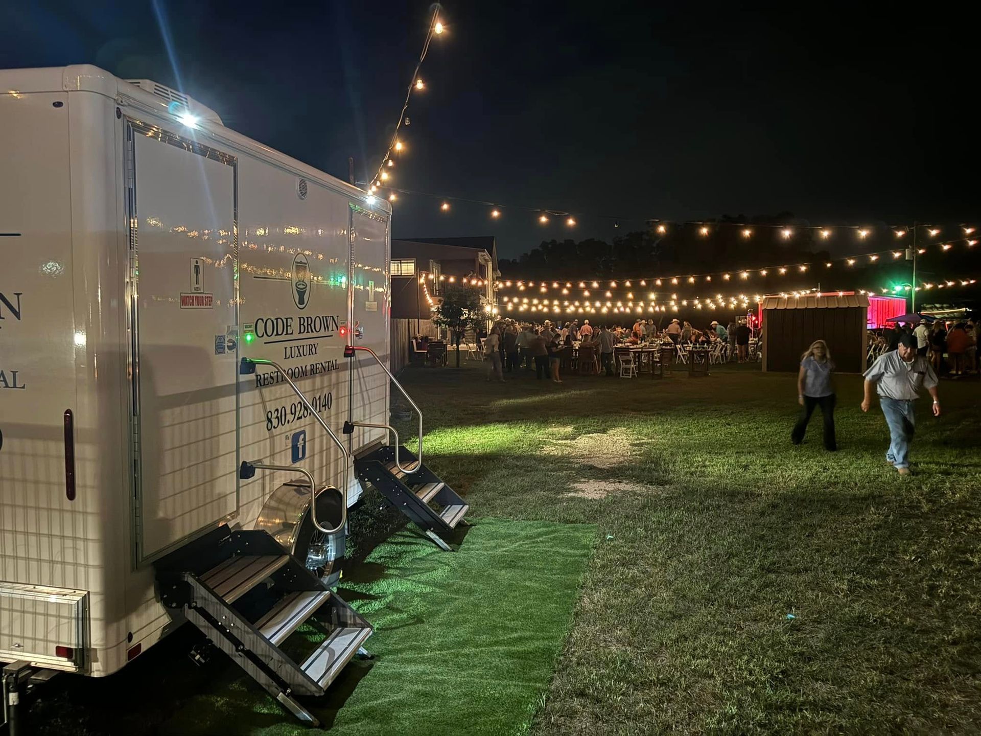 Nighttime outdoor event with string lights, a white trailer on the left, and people walking on a grassy field.