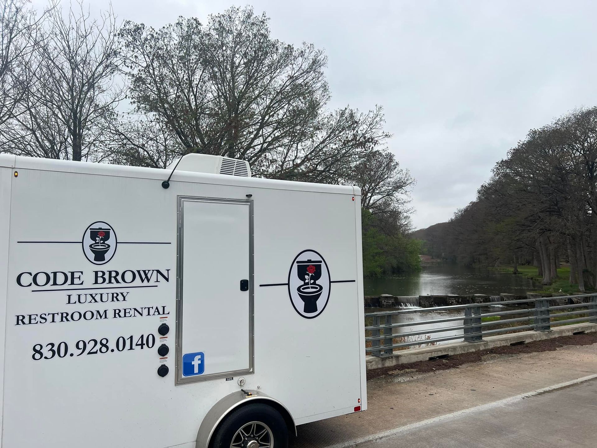 White Code Brown Luxury Restroom Rental trailer by a river, with trees and a bridge in the background