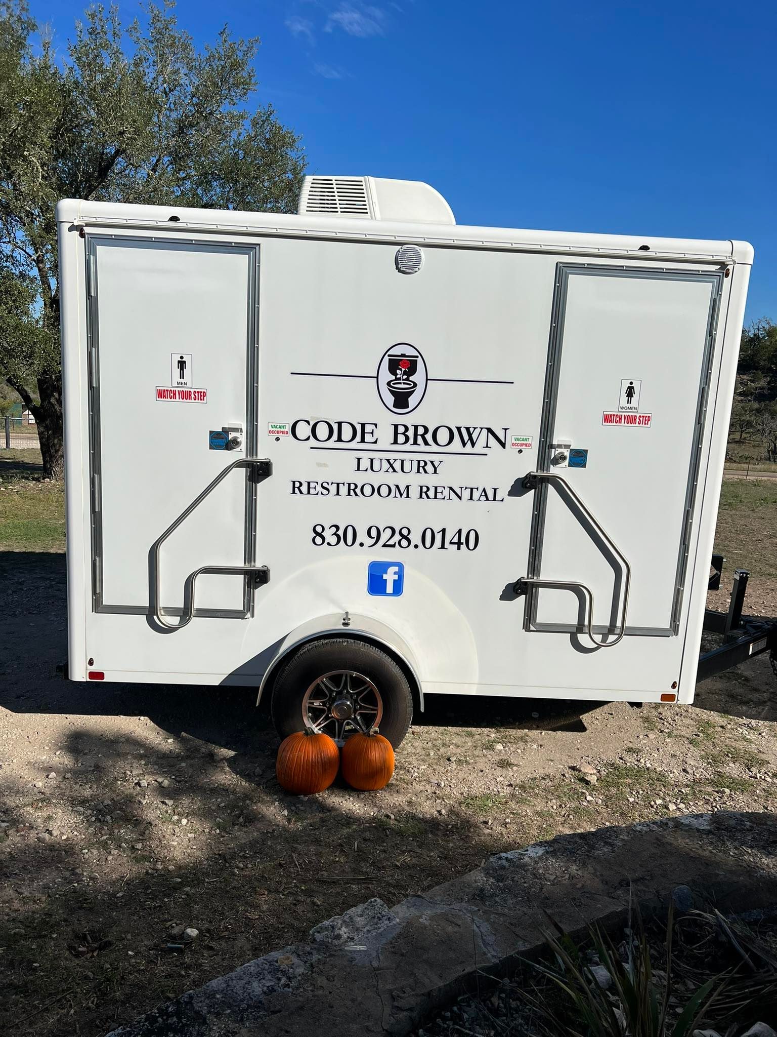 White food-truck trailer with logo and phone number, parked on gravel with pumpkins underneath.