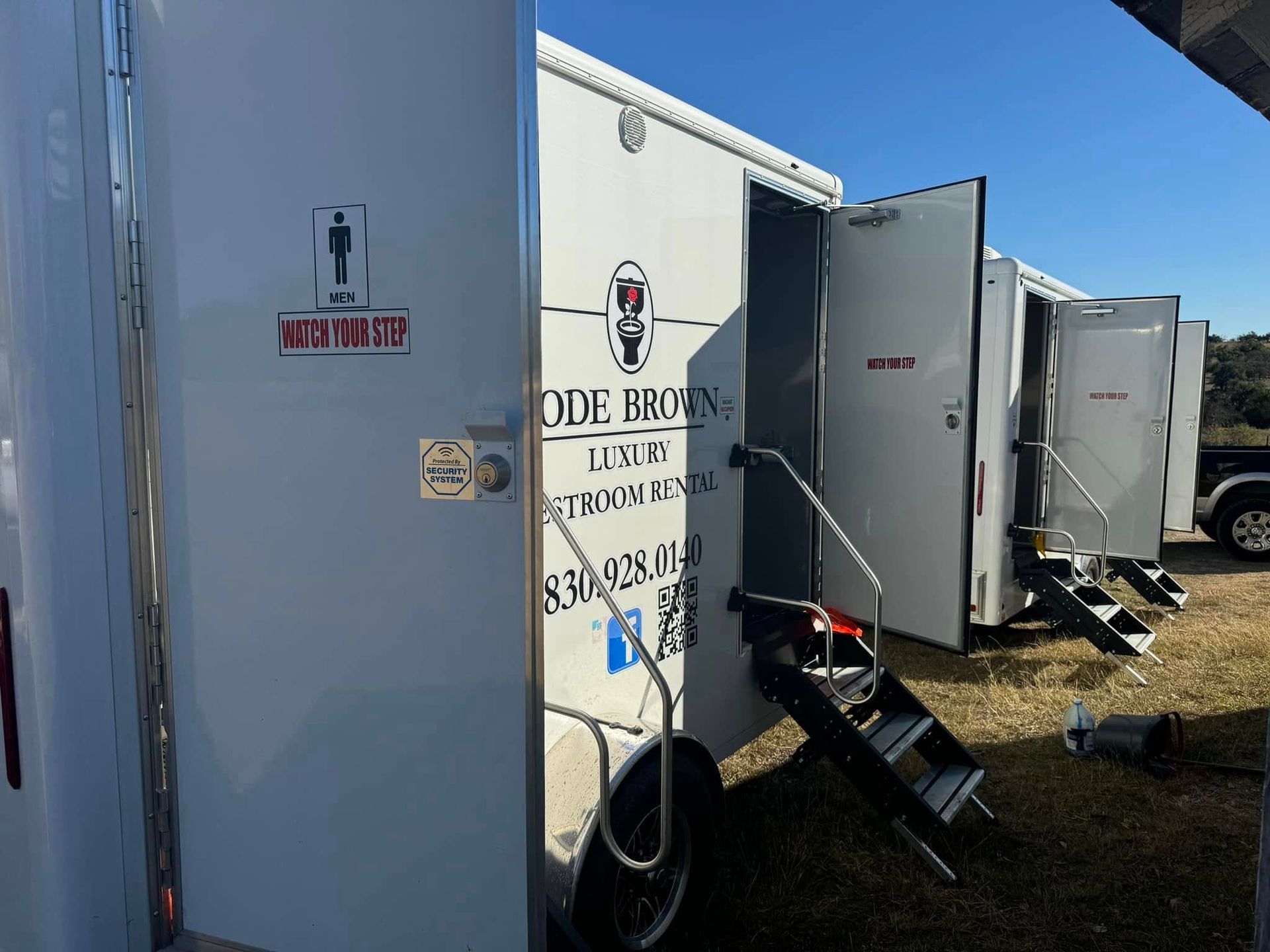 Row of gray portable restrooms and trailers on a grassy lot under a blue sky