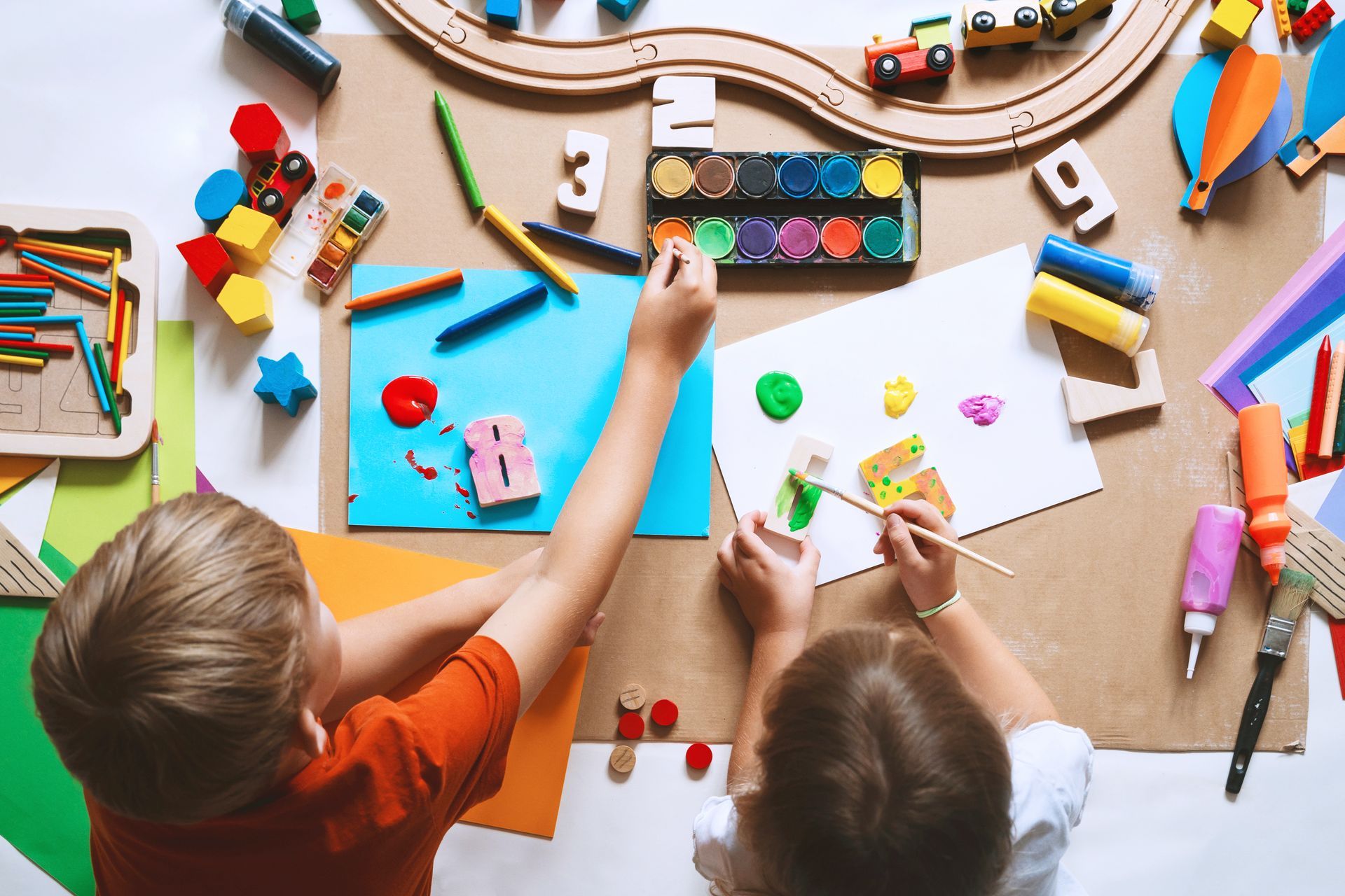 Children painting and playing with colorful art supplies and a wooden train track on a craft table
