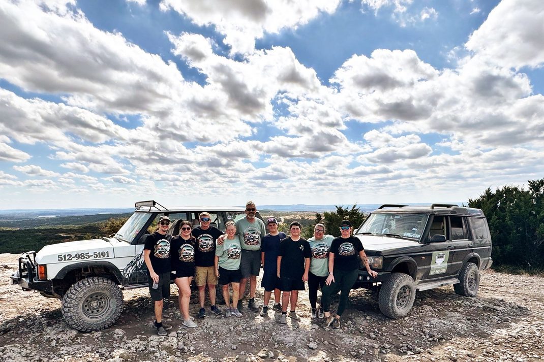 Group stands with off-road vehicles on a hilltop. Cloudy sky, two SUVs, and scenic overlook | Austin RoverWorks