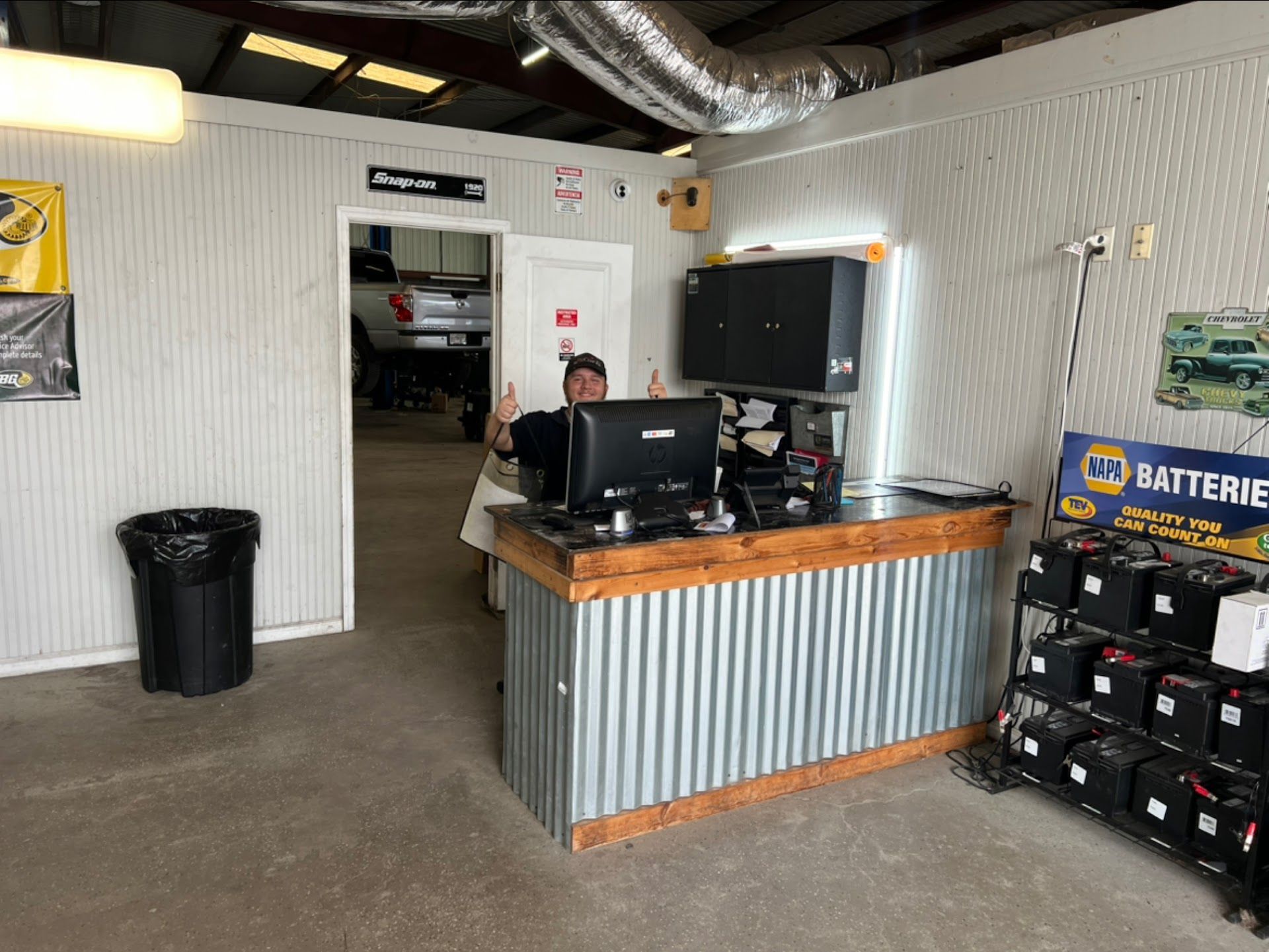 A person behind a front desk in an auto shop gives a thumbs-up. Corrugated metal and wooden trim on the desk. | CarDi Automotive