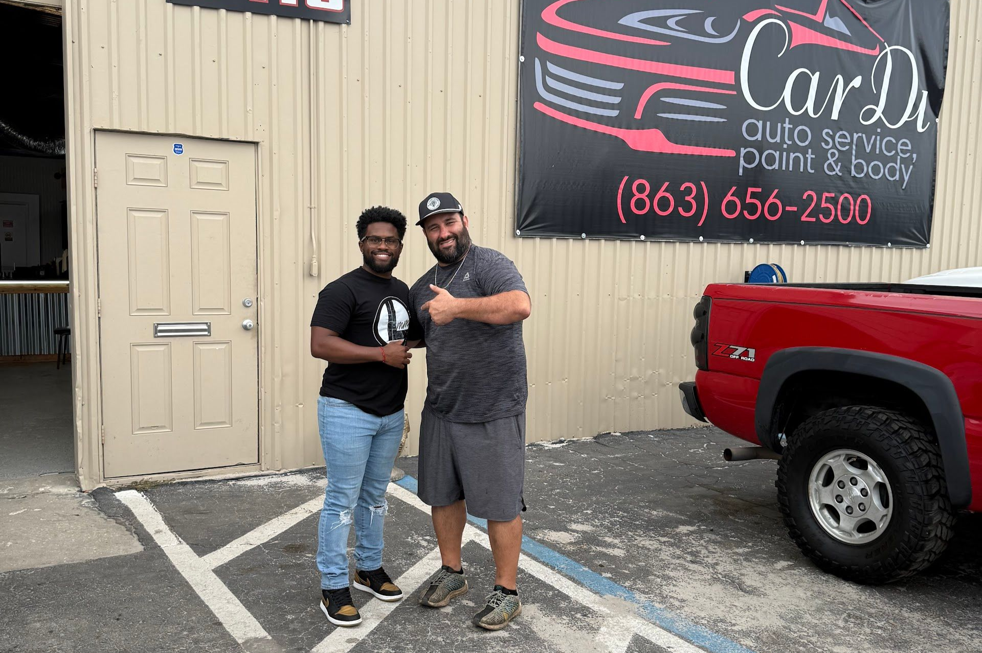 Two men standing in front of an auto shop, red truck visible. One gives a thumbs up. Shop sign with car graphic. | CarDi Automotive