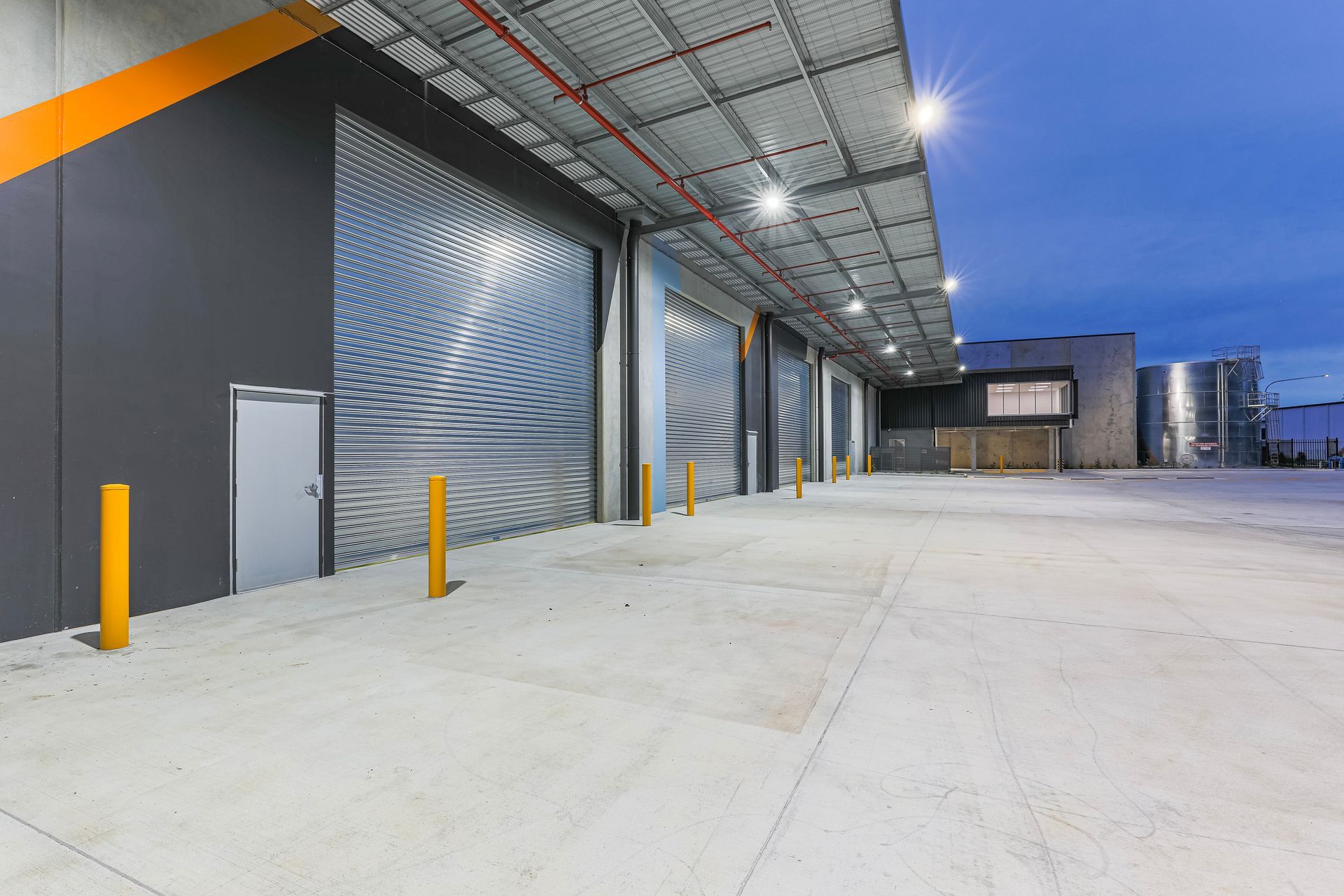 An industrial warehouse exterior at dusk, featuring multiple large gray metal roll-up doors and a concrete loading area. An industrial warehouse exterior at dusk, featuring multiple large gray metal roll-up doors and a concrete loading area.
