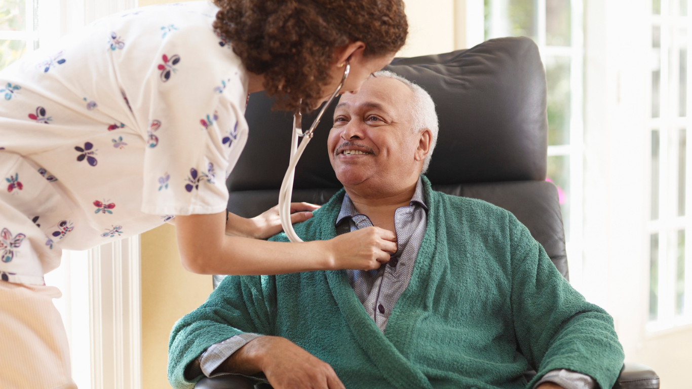 A healthcare worker listens to an elderly man's heart with a stethoscope in a sunlit room.