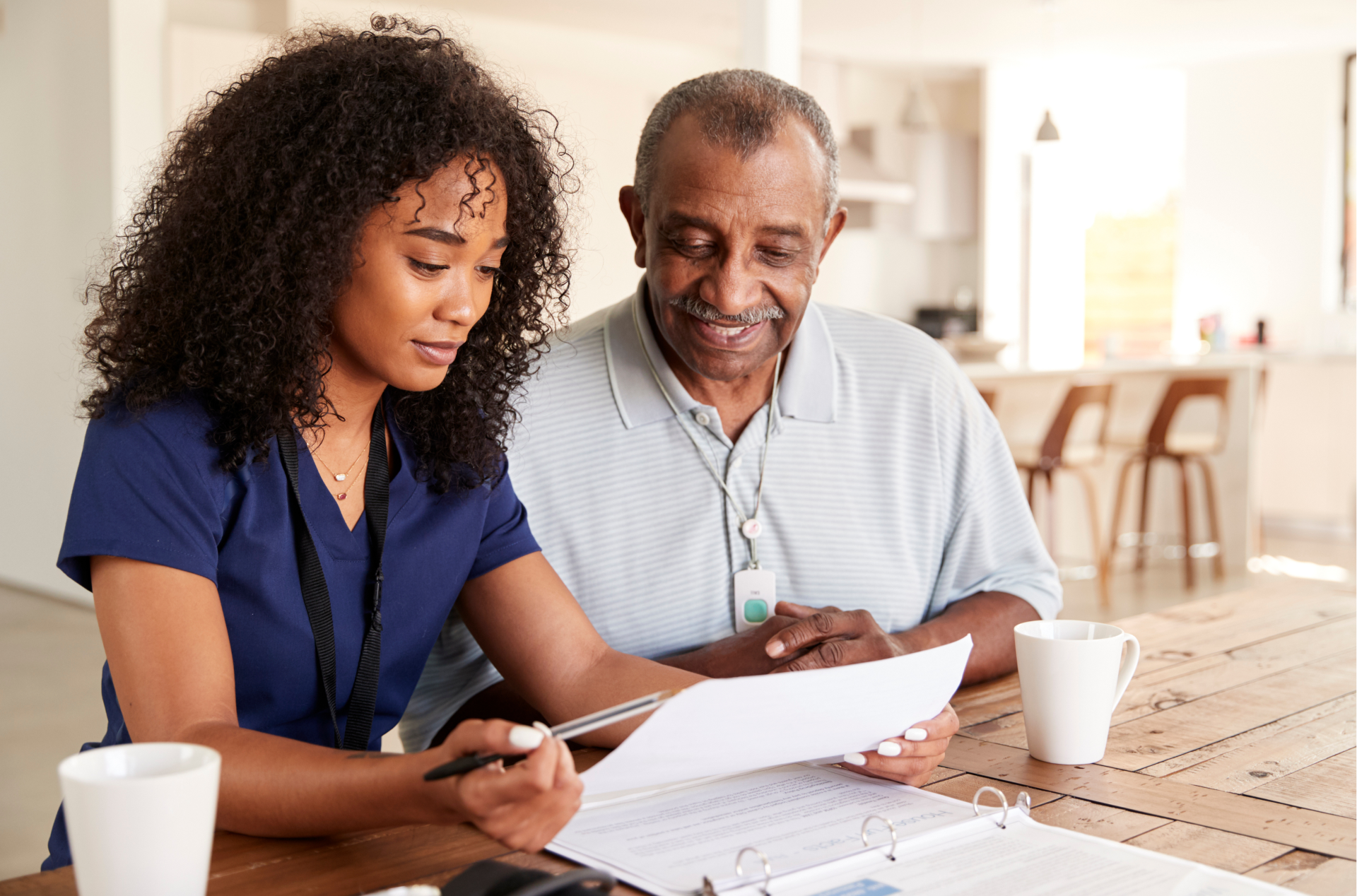 Caregiver in blue scrubs reviews paperwork with smiling senior man at a wooden table.