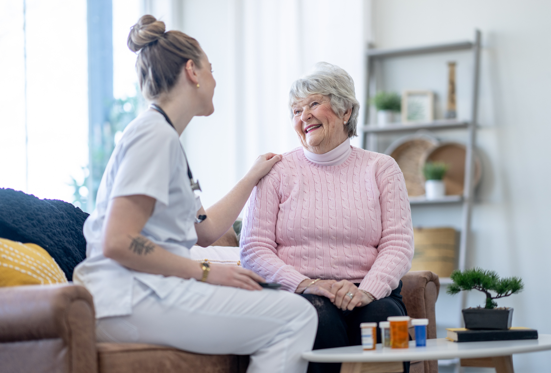 A young nurse with arm on shoulder of elderly woman, sitting and smiling. Living room.