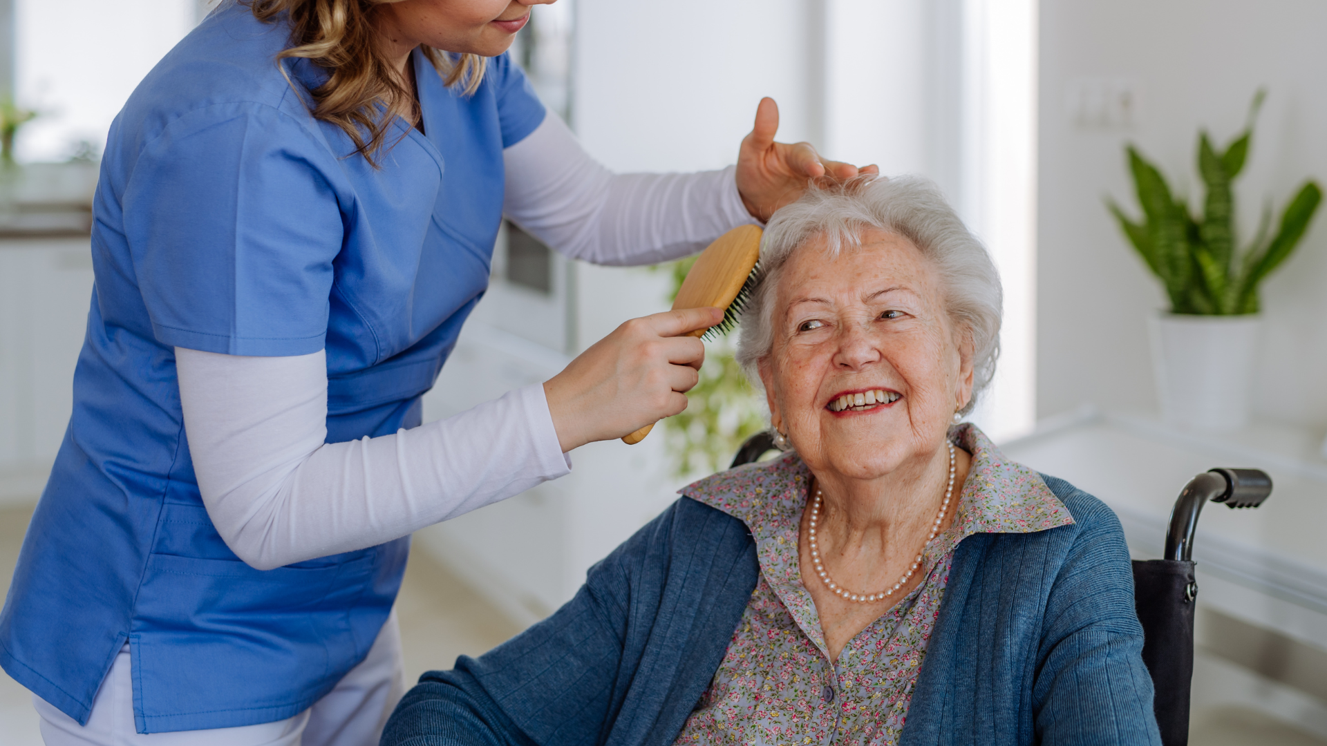 A healthcare worker brushes an elderly woman's hair; both smile. Indoors, sunny.