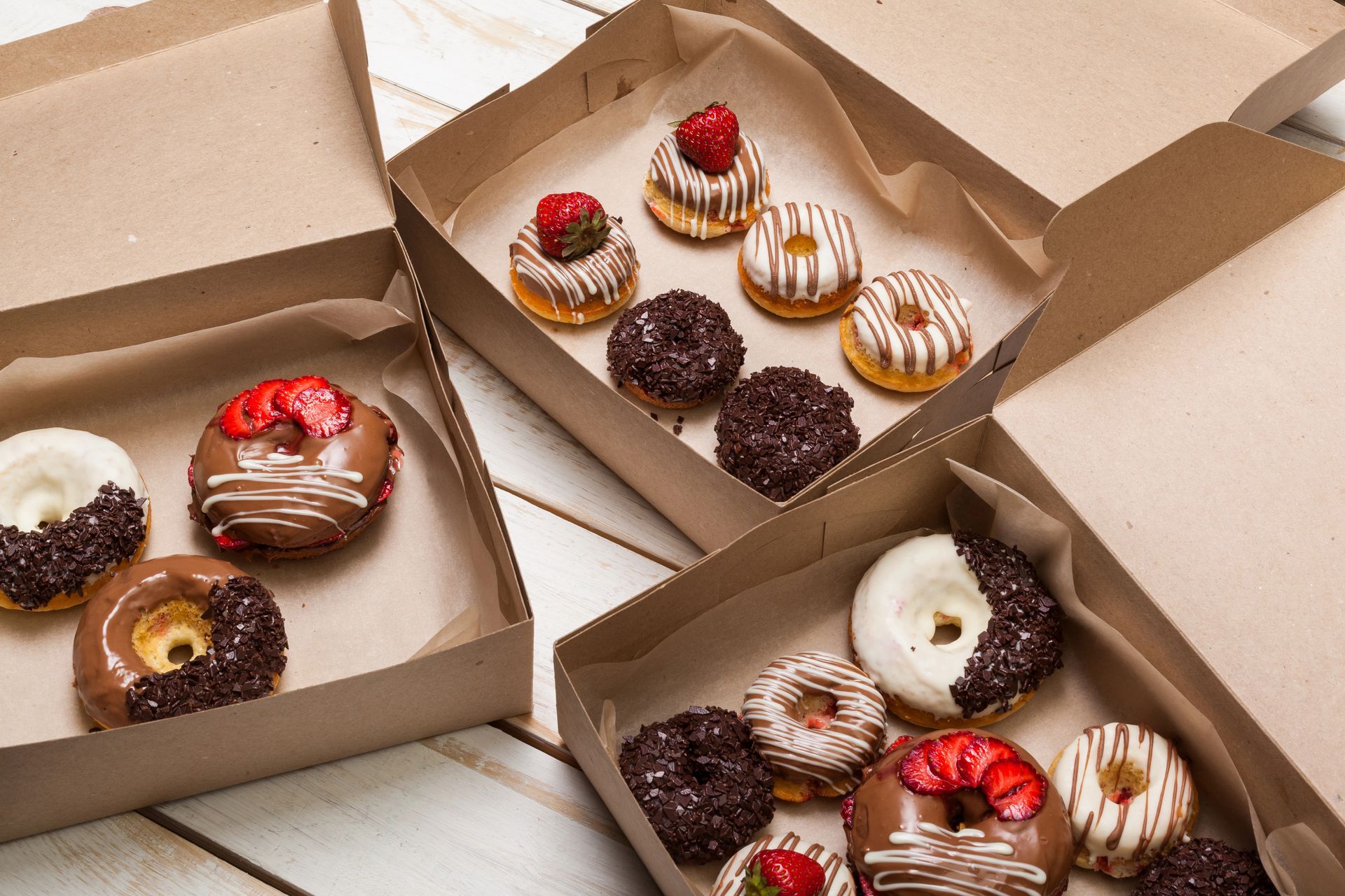 Donas en cajas de cartón abiertas; sabores surtidos con aderezos, dispuestas sobre una superficie de madera clara.