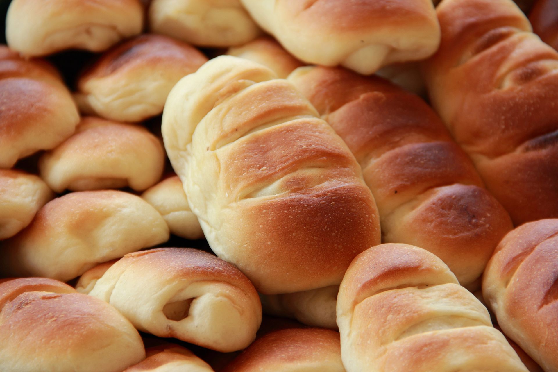 Primer plano de una pila de panecillos dorados recién horneados, de diversas formas, posiblemente en una panadería.
