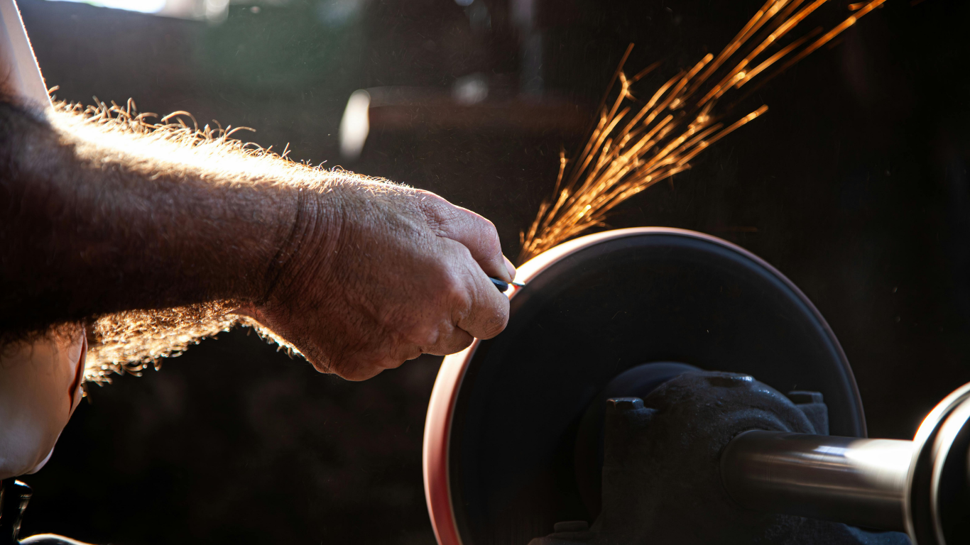 Person sharpening a metal object on a spinning grinding wheel, producing sparks in a dimly lit workshop.