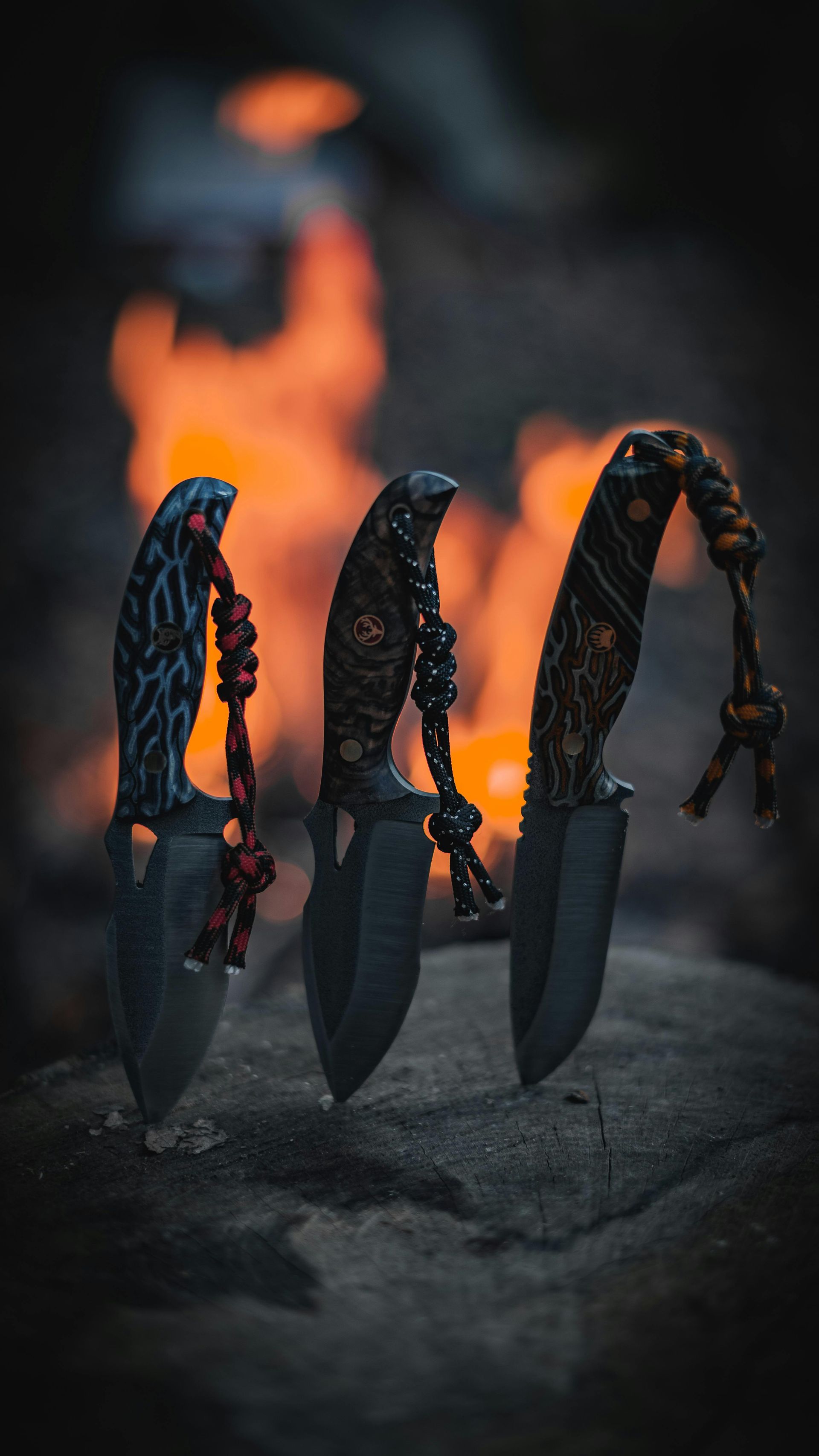 Three ornate knives with cord handles, set against a blurred campfire backdrop on a stone surface.
